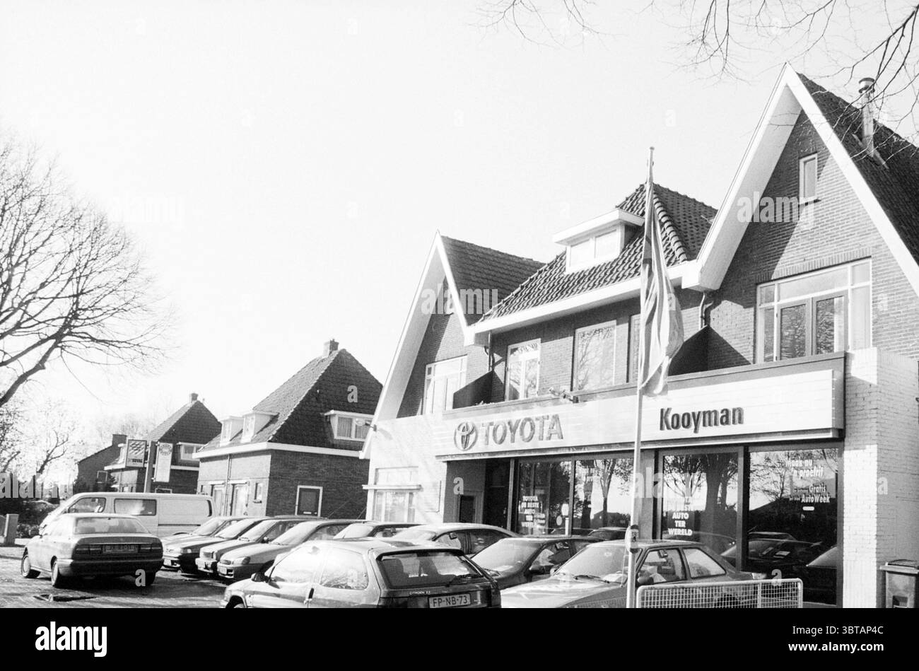 Garage Kooijman Vogelenzang Vogelenzang, Whizgle News, Dutch Desk, pays-Bas, 1950 - 2000 on 05-11-1996. Voici les rubriques de l'image. La scène capture un concessionnaire automobile placé contre un ciel clair et lumineux, suggérant une journée ensoleillée. Le bâtiment, une structure de deux étages, présente une combinaison de briques et de détails blancs, véhiculant une esthétique moderne mais accessible. Au premier plan, plusieurs voitures garées de différents modèles et couleurs créent un contraste vibrant, avec des nuances de noir, d'argent et de blanc prédominant. Leurs surfaces brillantes réfléchissent la lumière du soleil, améliorant l'impression de Br Banque D'Images
