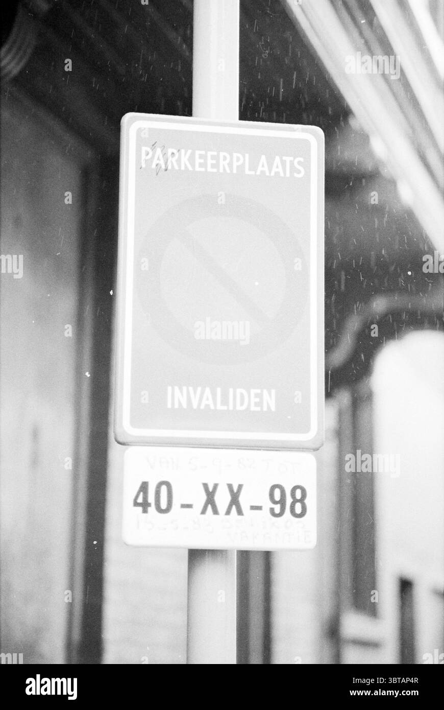 Panneau de stationnement pour handicapés avec plaque d'immatriculation, Whizgle News, Dutch Desk, pays-Bas, 1950 - 2000 on 16-11-1982. L'image inclut ces rubriques. La scène présente une composition monochromatique, caractérisée par des ombres douces et une atmosphère silencieuse. Un signe vertical domine le premier plan, affichant un emblème circulaire traversé par une ligne diagonale, indiquant des restrictions. La surface du panneau montre des signes d'usure et d'altération, ce qui ajoute de la texture à l'image globale. En arrière-plan, des caractéristiques architecturales peuvent être discernées, faisant allusion à un environnement urbain animé. La sortie Banque D'Images