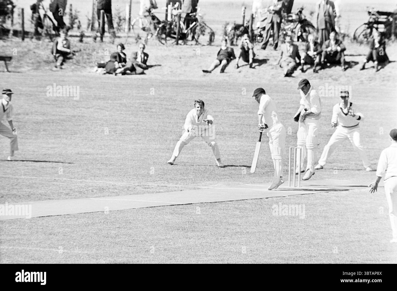 Haarlem - Red and White II Cricket, Whizgle News, Dutch Desk, pays-Bas, 1950 - 2000 le 24-06-1962. L'image contient ces rubriques. La scène représente un match de cricket animé se déroulant sur un vaste terrain vert. L'herbe est bien entretenue, vibrante et fraîchement coupée, offrant une excellente surface de jeu. Au premier plan, deux équipes sont engagées dans une compétition intense. D'un côté, un joueur de quilles est en cours de livraison, accroupi en avant avec une détermination focalisée. Il porte un uniforme de cricket blanc classique avec une casquette, soulignant la tenue traditionnelle du sport. Son expression véhicule la concentration, Banque D'Images
