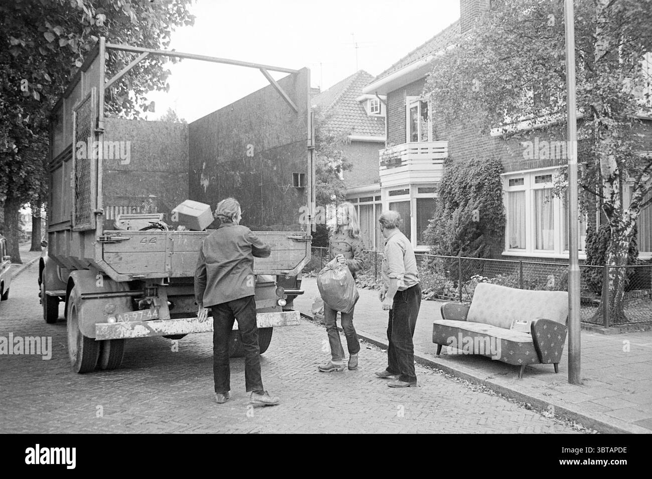 Déchets volumineux pour ordures ménagères, Whizgle News, Dutch Desk, pays-Bas, 1950 - 2000 sur 30-07-1973. L'image contient ces rubriques. Dans un cadre de rue tranquille, une scène se déroule avec un camion rustique stationné en angle le long du pavé. Le camion est vieilli et altéré, sa surface ornée de taches de saleté, laissant entendre son histoire industrieuse. Le dos ouvert révèle une cargaison, peut-être des sacs ou des outils, qui suggère qu'une journée de travail a commencé ou est en train de s'achever. Trois personnes sont engagées dans un effort de collaboration. Une personne, légèrement au premier plan, tient un grand sac ou Banque D'Images