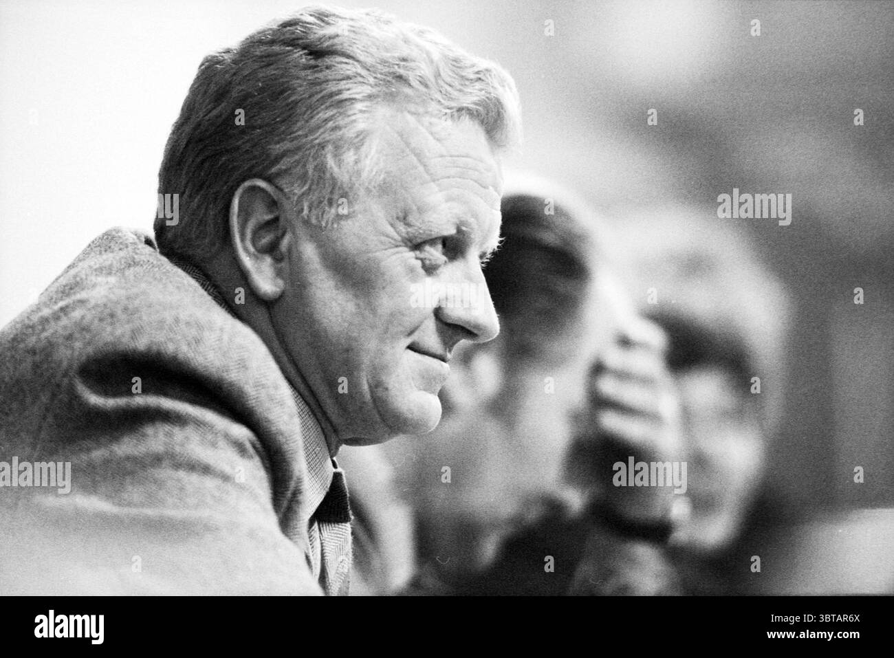 Débats budgétaires conseil de Haarlem conseil municipal de Haarlem Haarlem pays-Bas, Whizgle News, Dutch Desk, pays-Bas, 1950 - 2000 on 07-11-1979. L'image montre ces rubriques. Un homme est assis à une table, son profil est légèrement éclairé par la lumière ambiante. Ses cheveux, une nuance de gris clair, sont soigneusement peignés, révélant une expression composée et réfléchie. Il porte un costume sur mesure avec une texture subtile, probablement grise ou beige, complétant ses cheveux et ajoutant une touche de sophistication. L'arrière-plan présente des figures floues, suggérant un rassemblement ou une conversation entre plusieurs personnes. Le Overal Banque D'Images