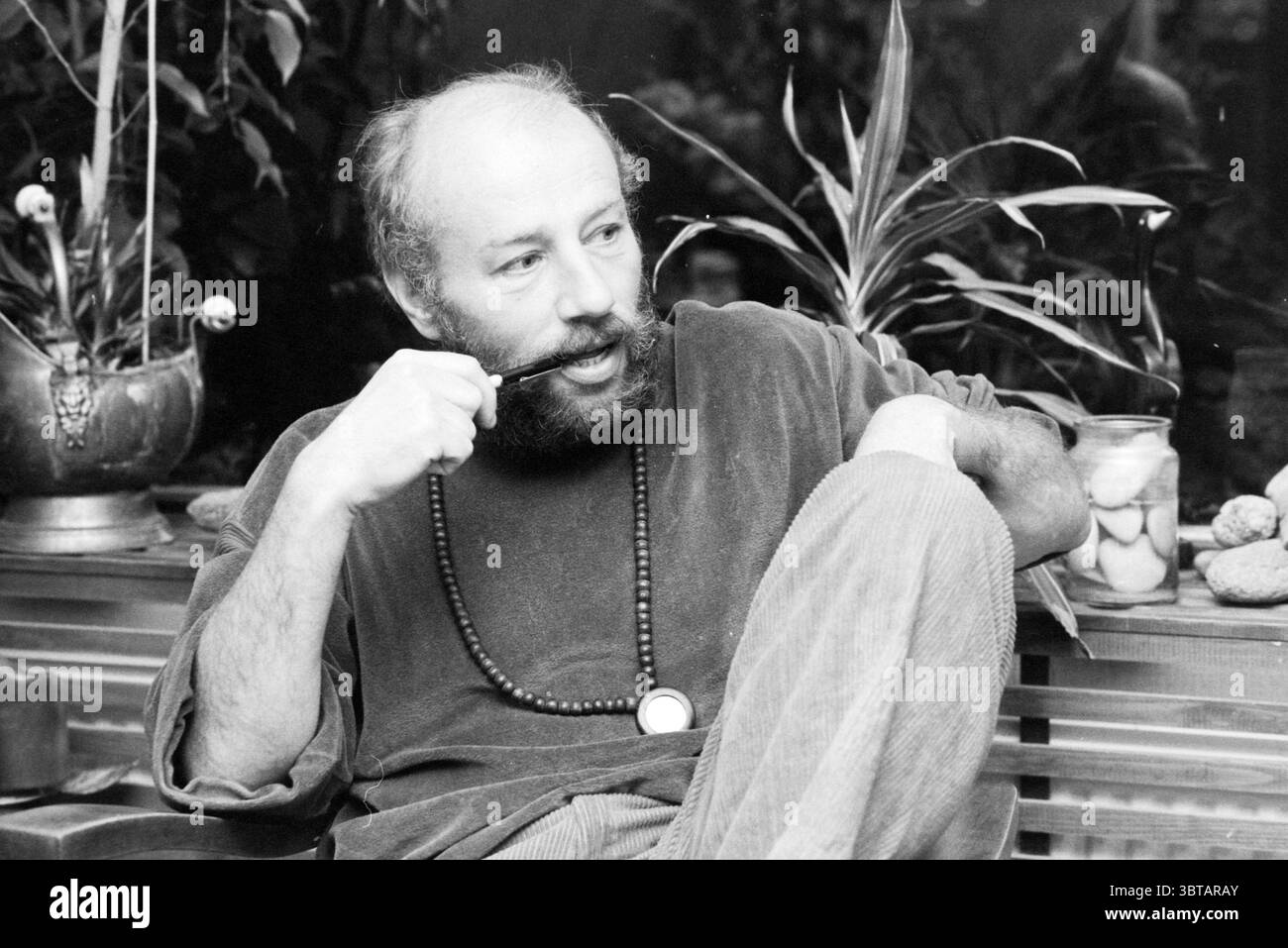 Portrait Mr. Van Gend Nieuw-Vennep Portraits Nieuw-Vennep, Whizgle News, Dutch Desk, pays-Bas, 1950 - 2000 le 24-04-1980. Voici les rubriques de l'image. La scène capture une figure contemplative assise dans une pose détendue. Il a une barbe pleine et tient soigneusement une pipe, suggérant un moment de réflexion. Sa tenue est décontractée et ample, avec des textures douces qui transmettent le confort. Autour de lui se trouvent des plantes vertes luxuriantes, améliorant l'ambiance naturelle du cadre. Les feuilles sont variées en forme et en taille, ajoutant profondeur et vitalité à la scène. Banque D'Images