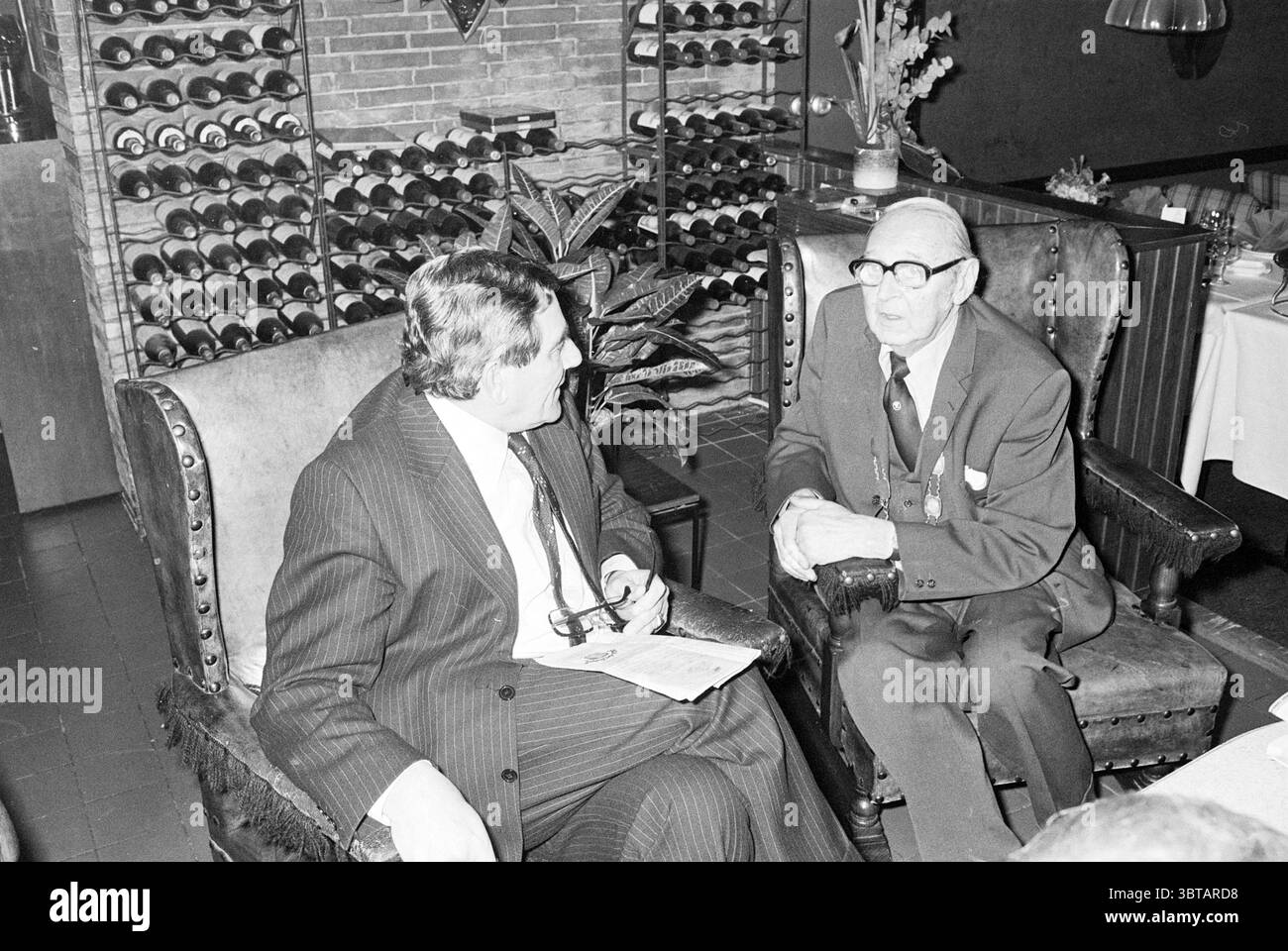 Déjeunez dans Learn Old Man Food and Eateries, Whizgle News, Dutch Desk, pays-Bas, 1950 - 2000 le 01-02-1979. Voici les rubriques de l'image. La scène met en scène deux hommes assis dans un cadre confortable et bien aménagé, caractérisé par une atmosphère chaleureuse et intime. Ils sont positionnés dans des fauteuils rembourrés encombrants qui ont un design vintage, soulignant un sentiment de confort et de conversation détendue. Le premier homme, vêtu d'un costume à rayures foncées, se penche légèrement en avant, suggérant un engagement et un intérêt pour la conversation. Il tient une paire de lunettes et semble curieux, son expr facial Banque D'Images