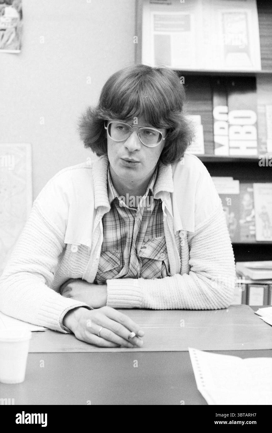 Carl de Jong 'Broadcast Piracy' Television, Whizgle News, Dutch Desk, pays-Bas, 1950 - 2000 le 26-05-1981. L'image inclut ces rubriques. La scène capture un jeune homme assis à une table, créant une atmosphère intime et introspective. Ses cheveux doux et brun clair tombent en vagues lâches, encadrant son visage, qui porte une expression contemplative. Il porte de grandes lunettes rondes qui ajoutent à son comportement réfléchi. Sa tenue se compose d'un cardigan de couleur claire superposé sur une chemise à carreaux, mettant en valeur un mélange de teintes douces comme le bleu et le vert. La texture du cardigan contraste avec Banque D'Images