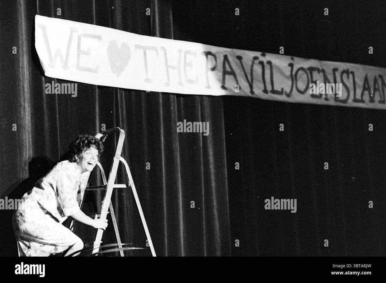 Soirée de fête Welgelegen Concertgebouw Foundation Parties Haarlem pays-Bas, Whizgle News, Dutch Desk, pays-Bas, 1950 - 2000 on 25-11-1986. Ces rubriques apparaissent dans l'image. Dans un auditorium faiblement éclairé, une femme est capturée en pleine action sur une échelle, son expression remplie de joie et d’enthousiasme. Elle a les cheveux bouclés, et sa tenue dispose d'une robe à motifs qui ajoute une touche de dynamisme au milieu de l'éclairage tamisé. La toile de fond est un rideau noir profond et riche qui encadre la scène, soulignant la présence de la femme au premier plan. Une grande bannière s'étend sur la partie supérieure du spa Banque D'Images