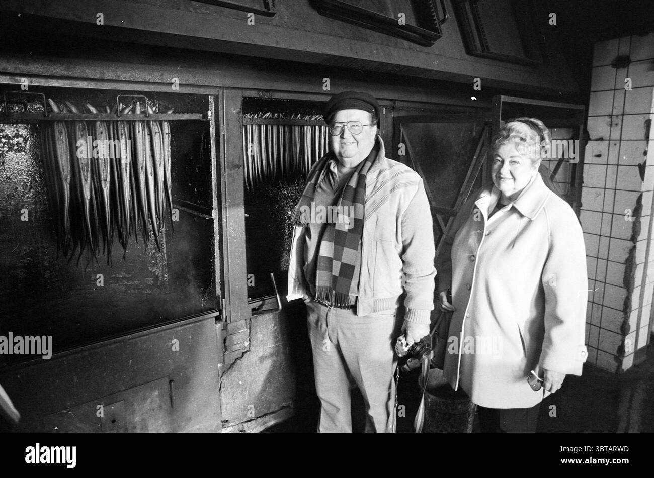 2 Américains de l'office de tourisme visitant Spaarndam étrangers Spaarndam, Whizgle News, Dutch Desk, pays-Bas, 1950 - 2000 on 07-12-1988. Ce sont les éléments de l'image. Dans un intérieur faiblement éclairé, deux personnes se tiennent côte à côte, dégageant toutes deux un sentiment de chaleur et de familiarité. L'homme, portant un béret foncé et des lunettes, est vêtu d'une écharpe à motifs et d'un pull léger, créant une apparence confortable et détendue. Son expression suggère un mélange d’amusement et de nostalgie. À côté de lui, la femme est habillée d'un manteau élégant et pâle qui contraste avec les tons plus sombres du décor. Elle Banque D'Images