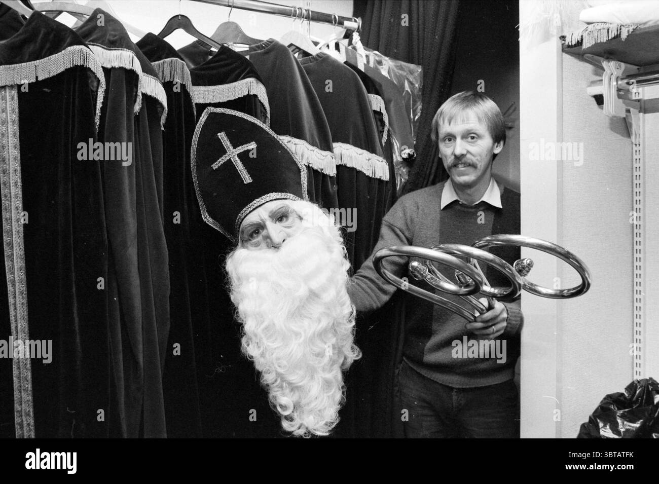 Jos 'Monnikkendam' with Sinterklaas Suits a obtenu Nicholas Sinterklaas, Whizgle News, Dutch Desk, The Netherlands, 1950 - 2000 on 03-12-1985. Ces rubriques sont illustrées dans l'image. Dans une zone de coulisses faiblement éclairée, l'atmosphère est à la fois intrigante et légèrement fantaisiste. Les costumes de velours sont suspendus à une étagère, leurs couleurs profondes et riches, principalement des rouges sombres et des noirs, contrastant avec le gris discret des murs. Un subtil éclat fait allusion au tissu luxueux, orné de bordures dorées ornées. Au premier plan, un homme se tient en partie caché derrière le rideau, tenant un grand masque élaboré avec un bea blanc Banque D'Images