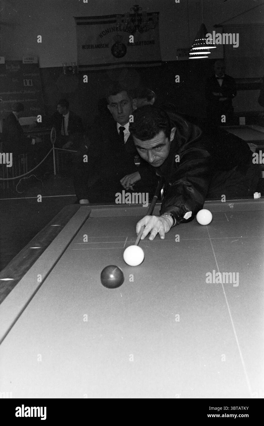 Joueur de billard néerlandais en action, Whizgle News, Dutch Desk, pays-Bas, 1950 - 2000 en 1966. Ce sont les éléments de l'image. Dans une pièce faiblement éclairée remplie d'un sentiment de concentration focalisée, deux hommes sont mis en évidence dans une scène engageante impliquant une table de billard. Le personnage principal, vêtu d'une veste sombre, se penche en avant avec une concentration intentionnelle, son front sillonné alors qu'il se prépare à frapper une balle blanche. Sa main, posée soigneusement sur le bord de la table, affiche un mélange de détermination et de finesse. L’atmosphère qui l’entoure est tendue, amplifiée par l’alose contrastée Banque D'Images
