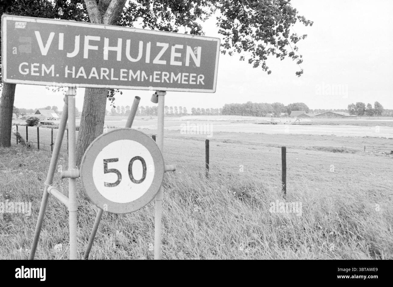 Sol de bulbe pulvérisé à Vijfhuizen Bloembollen Vijfhuizen, Whizgle News, Dutch Desk, pays-Bas, 1950 - 2000 le 31-07-1986. Ces rubriques apparaissent dans l'image. La scène présente un paysage rural caractérisé par une ambiance sereine et un peu discrète. Au premier plan, un panneau altéré se tient bien en évidence, délimitant la zone connue sous le nom de Vijfhuizen, accompagné d'un panneau de limite de vitesse circulaire indiquant 50. Les signes, avec leurs formes simples et leurs couleurs sourdes, sont placés sur un fond de verdure luxuriante. Le milieu environnant est marqué par de grands arbres, leurs feuilles formant une canopée dense ab Banque D'Images
