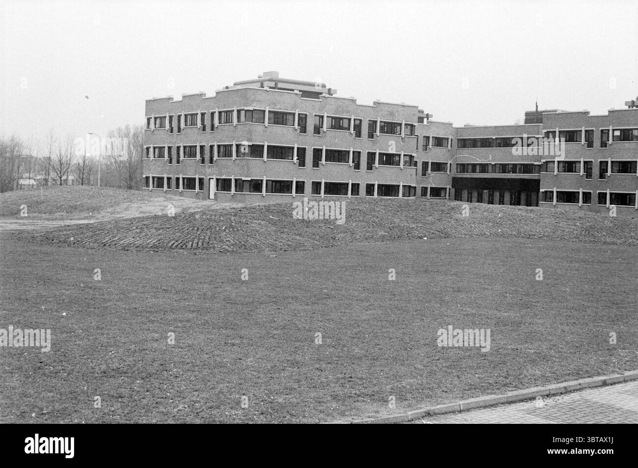 Zandbult derrière la mairie de Hoofddorp Zand Hoofddorp pays-Bas, Whizgle News, Dutch Desk, pays-Bas, 1950 - 2000 on 05-03-1984. L'image inclut ces rubriques. La scène représente une structure architecturale moderne caractérisée par son design angulaire et de nombreuses grandes fenêtres. Le bâtiment, construit principalement en briques, présente un motif répétitif qui ajoute du rythme à sa façade, avec des sections saillantes offrant une dynamique visuelle intéressante. La palette de couleurs est atténuée, dominée par des tons terreux de gris et de brun, ce qui donne au décor une qualité sombre et discrète. Dans le Banque D'Images