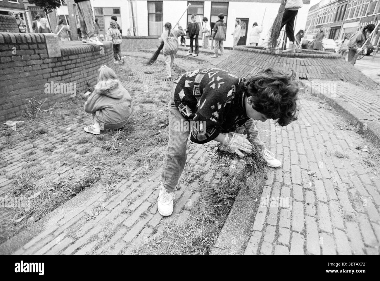 M. Schaffsen nettoie la place, Whizgle News, Dutch Desk, pays-Bas, 1950 - 2000 le 25-09-1991. L'image contient ces rubriques. Dans un milieu urbain animé, un groupe de personnes est engagé dans un effort de jardinage communautaire. Le sol est fait de briques rouges, créant un chemin distinct qui serpente à travers la scène. Plusieurs personnes peuvent être vues en train de travailler activement, certaines penchées vers le bas et tirant les mauvaises herbes de parcelles de sol fraîchement perturbé, tandis que d'autres se tiennent à proximité, surveillant la zone ou portant des outils. Les individus sont habillés dans des vêtements décontractés et confortables adaptés aux tâches extérieures, featur Banque D'Images