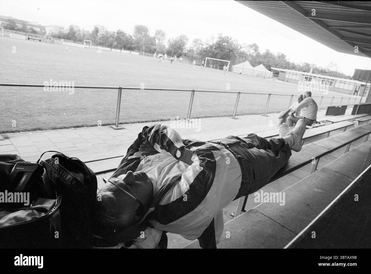 Entraîneur américain Atlanta, Whizgle News, Dutch Desk, pays-Bas, 1950 - 2000 sur 02-08-1992. Ces rubriques sont illustrées dans l'image. La scène capture un moment sur un terrain de sport, probablement un terrain de football, vu depuis un coin salon couvert. Au premier plan, deux individus occupent le banc. Une personne s'incline confortablement, vêtue d'une veste légère et d'un pantalon décontracté. Il a une posture détendue, peut-être se reposer après une longue journée. Sa tête est inclinée vers l'arrière, et des lunettes de soleil peuvent être vues perchées sur son visage, ajoutant une sensation de calme au cadre. A proximité, une autre figure est assise debout, engagée dans un plus actif Banque D'Images