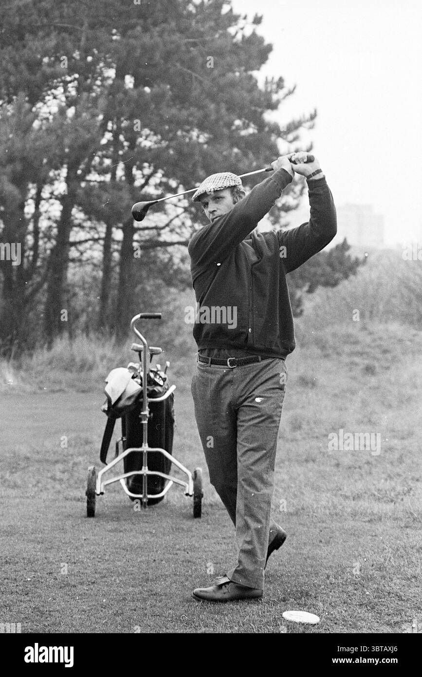 Barry Hughes sur le terrain de golf Golf Golf Football personnes et entraîneurs, Whizgle News, Dutch Desk, pays-Bas, 1950 - 2000 on 13-11-1975. Ces rubriques apparaissent dans l'image. Dans un cadre verdoyant et luxuriant, un homme est pris à mi-course, démontrant la concentration et la concentration. Il porte une casquette de style vintage avec un pull foncé et un pantalon ajusté, reflétant une tenue classique de golfeur. La palette de couleurs se compose de tons terreux, avec des verts riches de l'herbe et des arbres, contrastant avec ses vêtements plus sombres. Derrière lui, un sac de golf se trouve sur un petit chariot, faisant allusion aux outils du sport. Le sac est f Banque D'Images