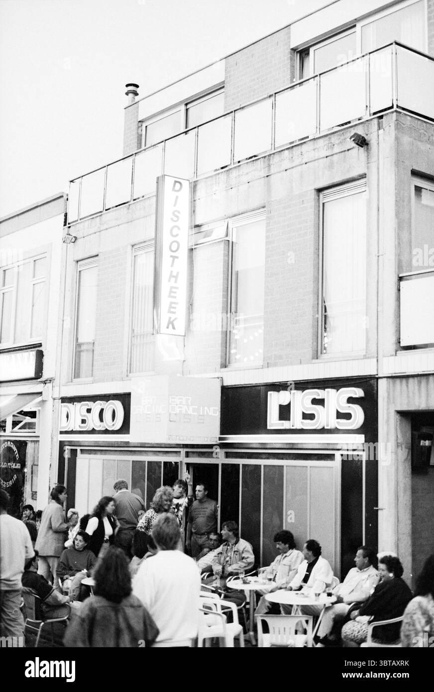 Nuit d'été à Zandvoort Zandvoort, Whizgle News, Dutch Desk, pays-Bas, 1950 - 2000 le 19-06-1992. L'image montre ces rubriques. La scène capture un rassemblement nocturne animé à l'extérieur d'un établissement dynamique. Le bâtiment, caractérisé par un style architectural moderne, dispose de grandes fenêtres qui reflètent la lumière douce et ambiante de l'intérieur. Sa façade est peinte dans des tons discrets, permettant aux enseignes au néon de sauter avec des couleurs vives, en particulier celle de la « DISCO » qui brille dans des teintes vives et électriques. Une foule de personnes diverses est réunie à l'extérieur, créant une atmosphère dynamique. Ils sont habillés Banque D'Images