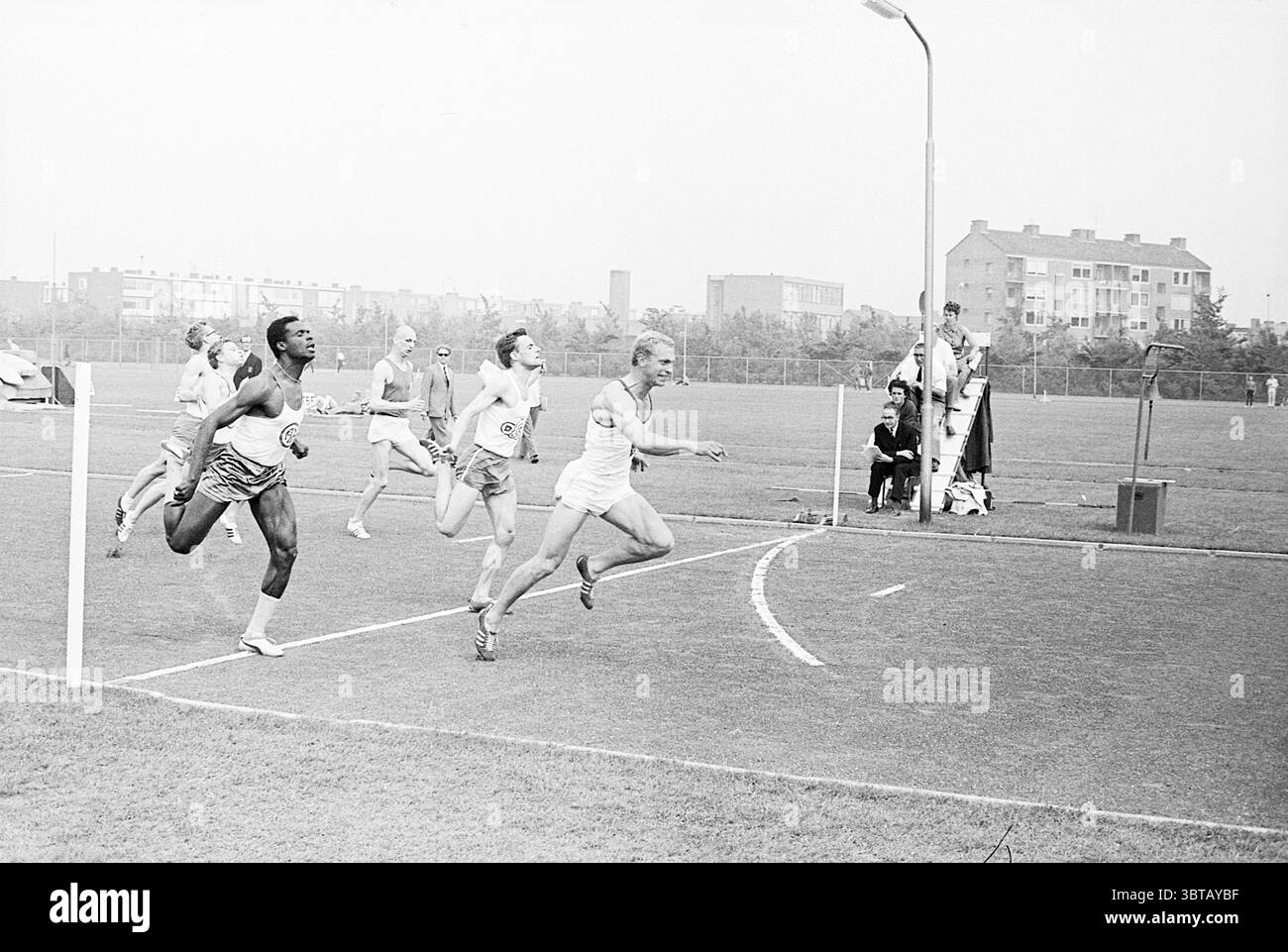 Compétitions d'athlétisme., Whizgle News, Dutch Desk, pays-Bas, 1950 - 2000. L'image montre ces rubriques. La scène capture un moment dynamique lors d'un événement de course sur un terrain de sport spacieux. Au premier plan, un groupe de quatre coureurs masculins est représenté en sprint complet, leurs corps inclinés vers l'avant, montrant une concentration et une détermination intenses. Les athlètes sont habillés dans différents styles d'équipement sportif : l'un est en short rayé et un haut sans manches, tandis qu'un autre arbore un uniforme blanc léger, permettant la liberté de mouvement. L'arrière-plan présente une large étendue de tur vert Banque D'Images