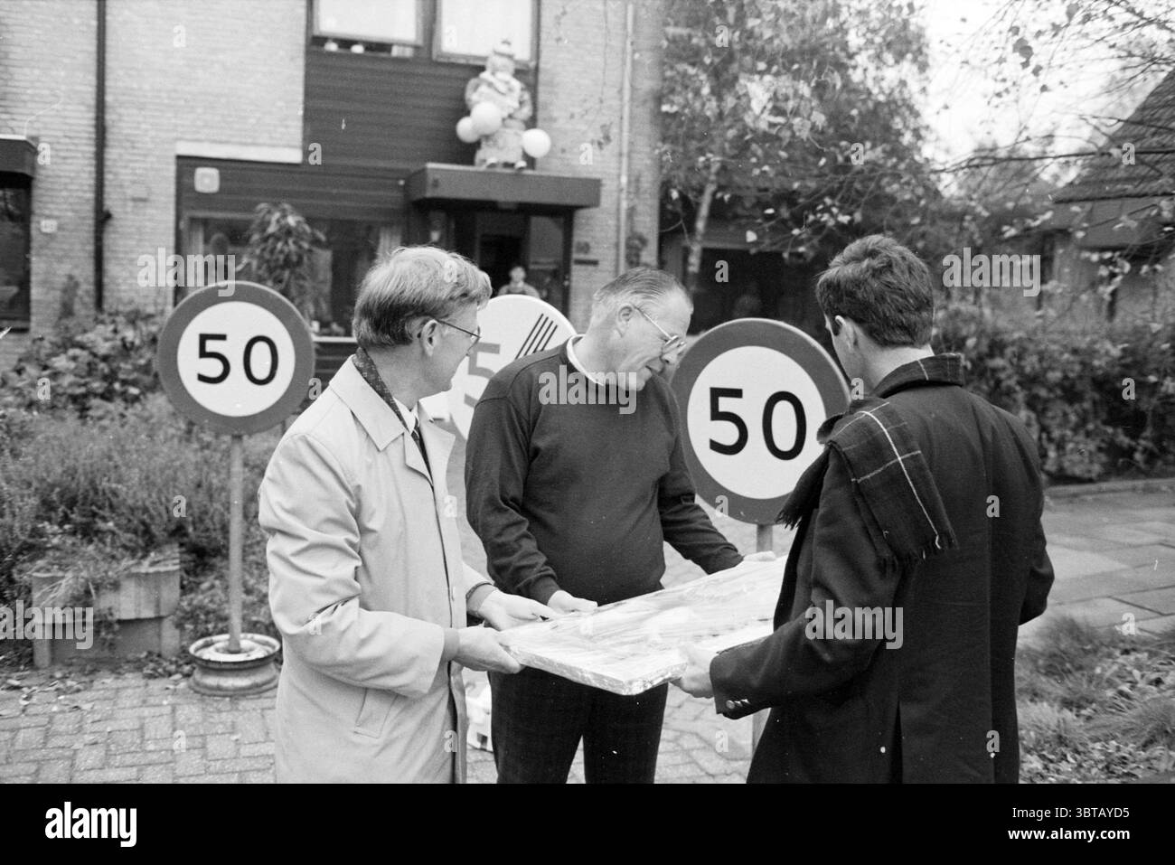 Alderman Auer Hoofddorp 50 ans anniversaires Aldermen Hoofddorp pays-Bas, Whizgle News, Dutch Desk, pays-Bas, 1950 - 2000 on 11-10-1988. Ce sont les éléments de l'image. En banlieue, trois personnes discutent près d'une paire de panneaux circulaires affichant le chiffre « 50 », indiquant une limite de vitesse. Les panneaux sont placés en évidence de chaque côté de la scène, entourés de verdure et d'arbustes doux, suggérant un environnement bien entretenu. Deux hommes sont positionnés plus près du premier plan ; l'un porte des lunettes et un trench-coat de couleur claire, tandis que th Banque D'Images
