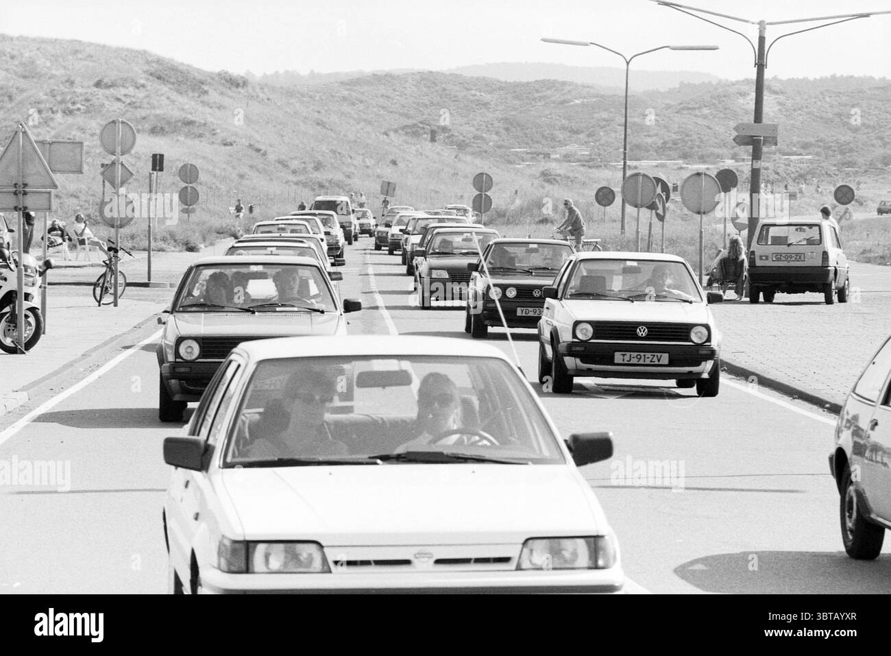 Les automobilistes sur le Zeeweg maintenus par le Haarlem Mosquito Round. Bloemendaal Zeeweg, Whizgle News, Dutch Desk, pays-Bas, 1950 - 2000 le 02-08-1990. Ces rubriques apparaissent dans l'image. La scène capture une route très fréquentée, animée de véhicules alignés, créant un sentiment de mouvement et d'urgence. Les voitures varient en taille et en style, principalement des modèles plus anciens caractérisés par leurs designs classiques et leurs courbes subtiles. Certains sont peints dans des couleurs discrètes comme le blanc, le gris et le noir, contribuant à une sensation intemporelle, tandis que d'autres introduisent occasionnellement des éclaboussures de couleur. Autour des voitures se trouve un v Banque D'Images