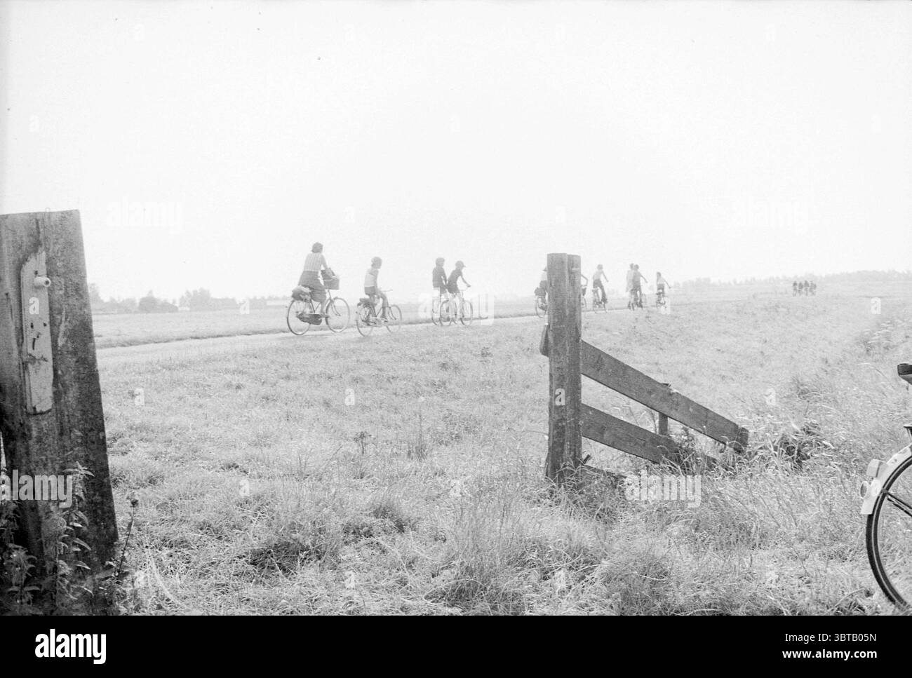 Événement cycliste de quatre jours Heemskerk Cycling pistes cyclables cyclistes, Whizgle News, Dutch Desk, pays-Bas, 1950 - 2000 le 26-07-1978. Voici les rubriques de l'image. La scène présente un paysage serein et pastoral où un groupe de cyclistes traverse un long chemin sinueux qui traverse un vaste champ. Les tons doux et discrets de noir et blanc créent une atmosphère nostalgique, évoquant un sentiment de tranquillité et de simplicité. À gauche, une porte en bois altérée se trouve légèrement entrouverte, sa texture rustique complétant l'environnement naturel. Les poteaux de la porte sont grossièrement taillés, indiquant l'âge et un conn Banque D'Images
