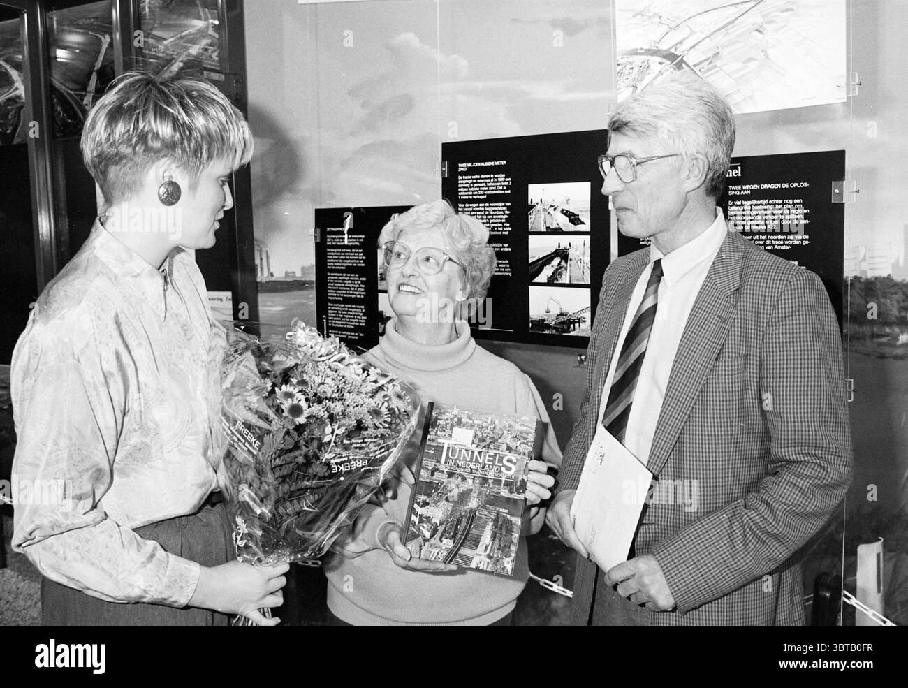 10000ème visiteur du centre d'information Wijkertunnel, Whizgle News, Dutch Desk, pays-Bas, 1950 - 2000 sur 19-04-1994. Voici les rubriques de l'image. Dans un cadre intérieur bien éclairé, trois individus sont réunis en groupe étroit, créant une atmosphère chaleureuse et intime. Une femme à gauche, aux cheveux courts blonds platine, porte de grandes boucles d'oreilles et une tenue élégante, suggérant une sensibilité moderne. Elle tient un bouquet rempli de fleurs vibrantes, mettant en valeur des teintes de rose, jaune et blanc, qui ajoute une touche de fraîcheur à la scène. Au centre, une femme plus âgée avec un h court et blanc Banque D'Images