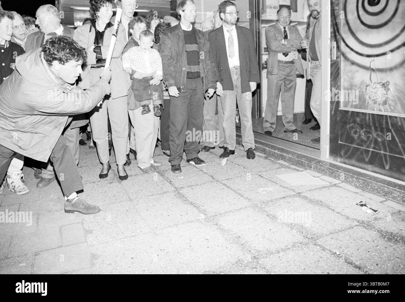 Réouverture Korstanje Santpoort-Noord ouverture ouvertures mises en service Santpoort-Noord, Whizgle News, Dutch Desk, pays-Bas, 1950 - 2000 on 07-03-1990. Ces rubriques apparaissent dans l'image. La scène se déroule la nuit dans une zone urbaine, éclairée par des lumières artificielles provenant de fenêtres et de lampadaires à proximité. Un groupe diversifié de personnes se réunit sur un trottoir pavé, la plupart d'entre eux faisant face à un point d'intérêt central juste devant. Au premier plan, une personne s'accroupit, intensément concentrée sur le sol. Ils sont habillés de façon décontractée, avec des vêtements plus sombres contrastant avec les pierres plus claires du trottoir. Thei Banque D'Images