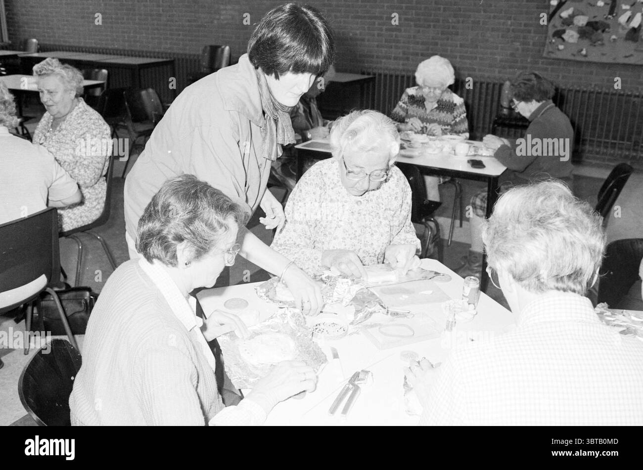Les femmes de pays fabriquent des objets à partir de Dough Hobby Women, Whizgle News, Dutch Desk, pays-Bas, 1950 - 2000 sur 13-11-1984. Ces rubriques sont illustrées dans l'image. La scène capture un cadre intérieur animé, probablement un centre communautaire ou une salle d'activités, caractérisé par une atmosphère chaleureuse et accueillante. La salle est remplie de diverses tables rondes, chacune entourée de personnes âgées engagées dans une activité artisanale ou culinaire. Au premier plan, un groupe de quatre femmes âgées se concentre sur leurs tâches, avec leur attention attirée sur une table aménagée avec des matériaux d'artisanat. Les femmes sont habillées en confortable, patt Banque D'Images