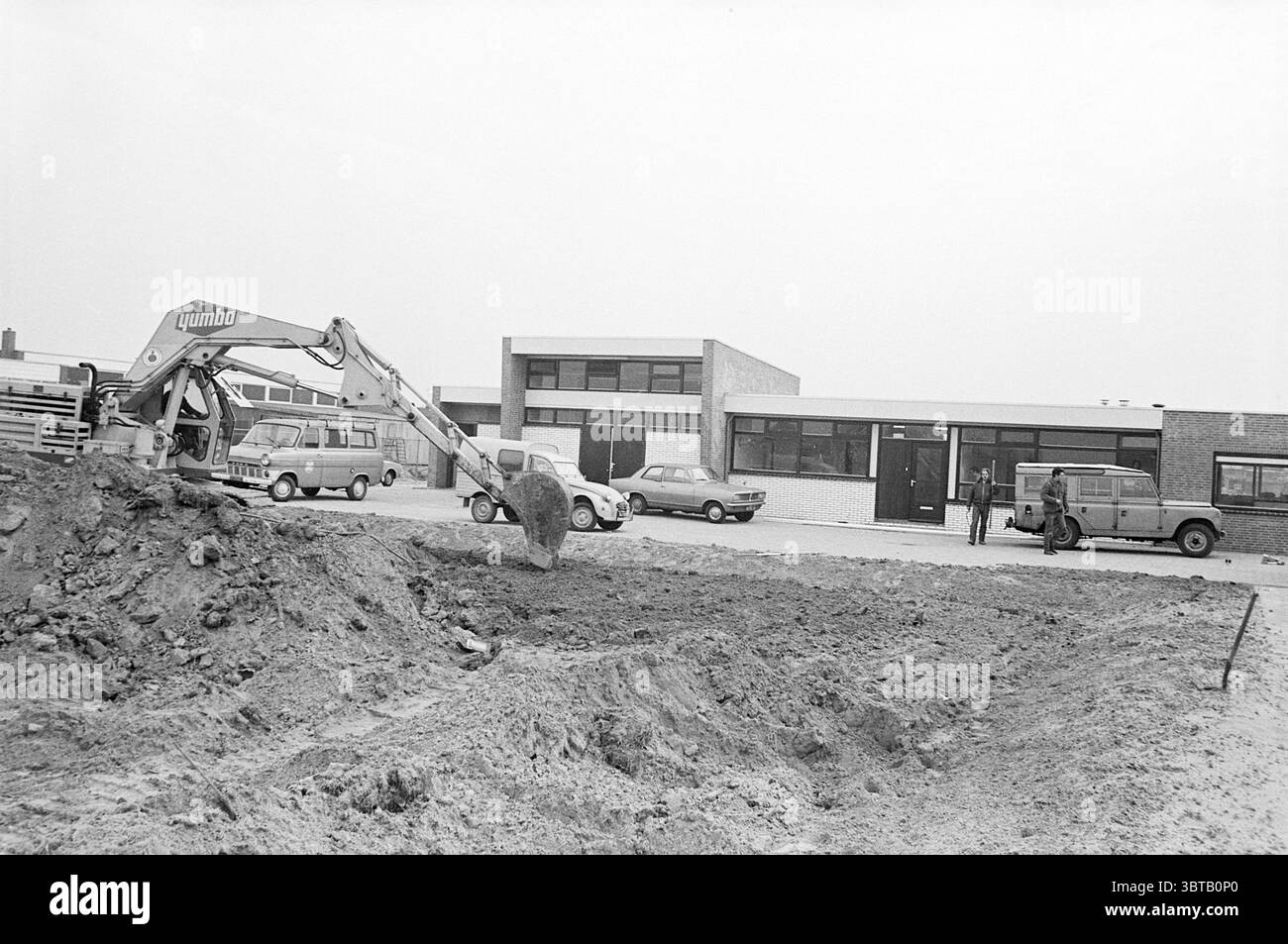Ext. Meat inspection circle building Exterior, Whizgle News, Dutch Desk, pays-Bas, 1950 - 2000 on 18-01-1972. Ces rubriques apparaissent dans l'image. Dans la scène, un chantier de construction est mis en évidence, caractérisé par un grand monticule de terre et de sable qui a été fraîchement excavé. Une pelle hydraulique, avec son bras articulé distinctif relevé haut, est engagée dans le creusement, son godet dirigé vers le sol, créant une impression de travail actif. À gauche, plusieurs véhicules stationnés sont alignés, avec une combinaison de modèles plus anciens avec différentes nuances de gris et de beige, contribuant à Banque D'Images