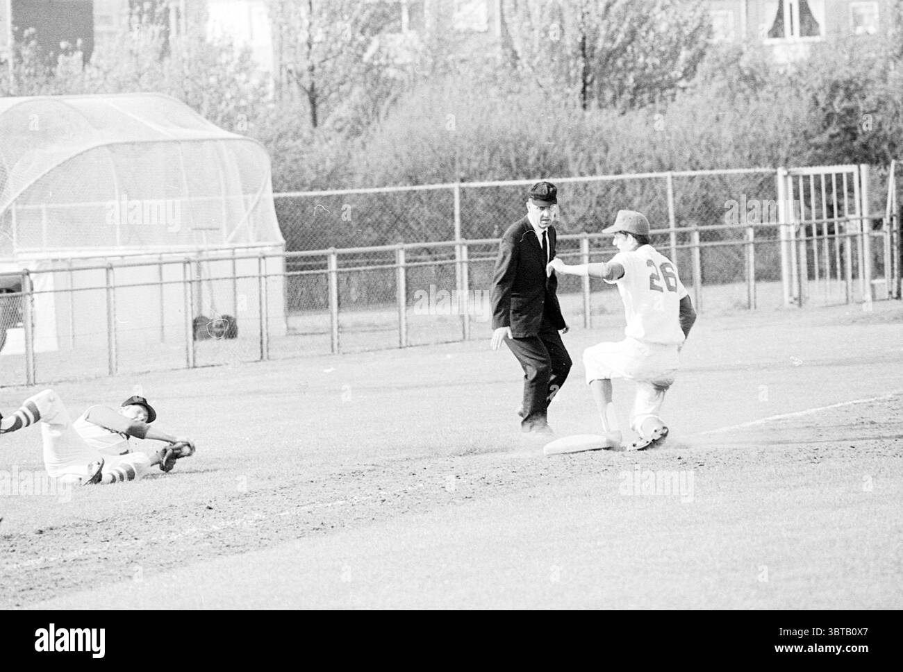 Haarlem Nicols - Feijenoord Baseball, Whizgle News, Dutch Desk, pays-Bas, 1950 - 2000 le 11-05-1969. L'image inclut ces rubriques. La scène se déroule sur un terrain de baseball herbeux, baigné dans une palette monochrome, principalement avec des nuances de gris. Deux joueurs sont au centre de l'action : l'un glisse dans une base, paraissant aéroporté avec sa jambe gauche tendue, tandis que son bras droit appuie contre le sol pour le soutenir. Son uniforme est marqué avec le numéro '26' au dos, faisant allusion à son rôle dans l'équipe. A proximité, un arbitre, vêtu d'une tenue traditionnelle avec une casquette, se tient dans un commandement Banque D'Images