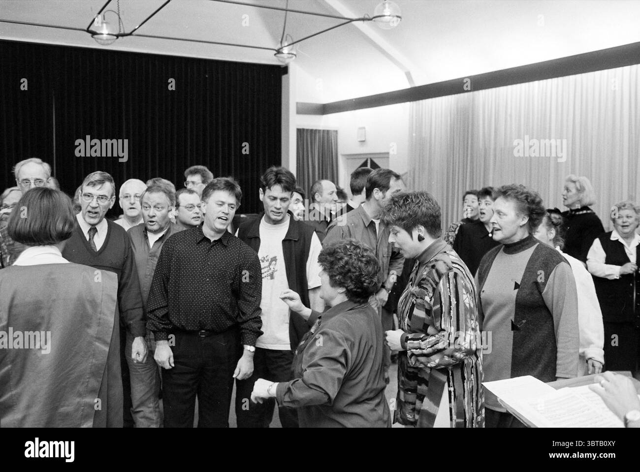 Association de répétition d'opérette vides IJm. IJmuiden pays-Bas, Whizgle News, Dutch Desk, pays-Bas, 1950 - 2000 le 09-04-1995. L'image contient ces rubriques. Dans un rassemblement animé, un groupe diversifié d'individus se tient étroitement ensemble, engagés dans une interaction énergique. La scène est vivante avec un sentiment de camaraderie et d'enthousiasme. Des gens de divers âges et origines sont présents ; certains sont en pleine chanson, leurs expressions animées et joyeuses, tandis que d'autres écoutent attentivement, hochant la tête en rythme. Les individus ont des coiffures et des styles de vêtements variés, ajoutant à l'atmosphère dynamique. Banque D'Images