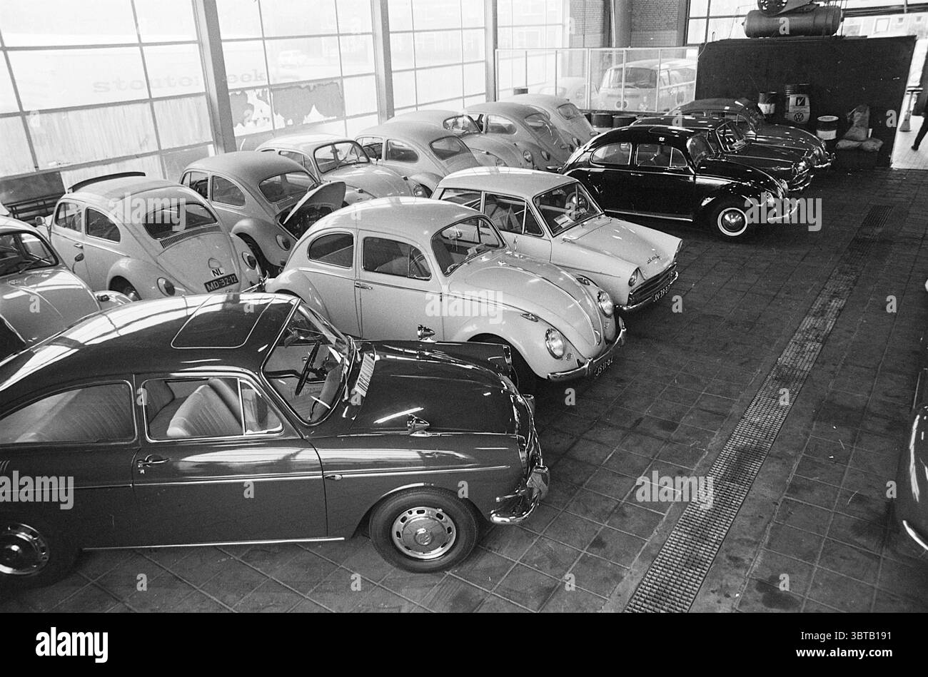 Report garage de Boer Report, Whizgle News, Dutch Desk, pays-Bas, 1950 - 2000 sur 07-10-1967. Ces rubriques sont illustrées dans l'image. La scène capture une salle d'exposition automobile vintage, caractérisée par un intérieur spacieux baigné d'une lumière douce et diffuse filtrant à travers de grandes fenêtres. L’ambiance est nostalgique, avec une ambiance industrielle subtile. Au premier plan, une série de voitures classiques sont disposées en rangées soignées, chacune présentant des designs distinctifs d'une époque révolue. Les véhicules sont principalement une palette de tons discrets : blancs doux, noirs profonds et nuances de gris. Une voiture se démarque avec un Li Banque D'Images
