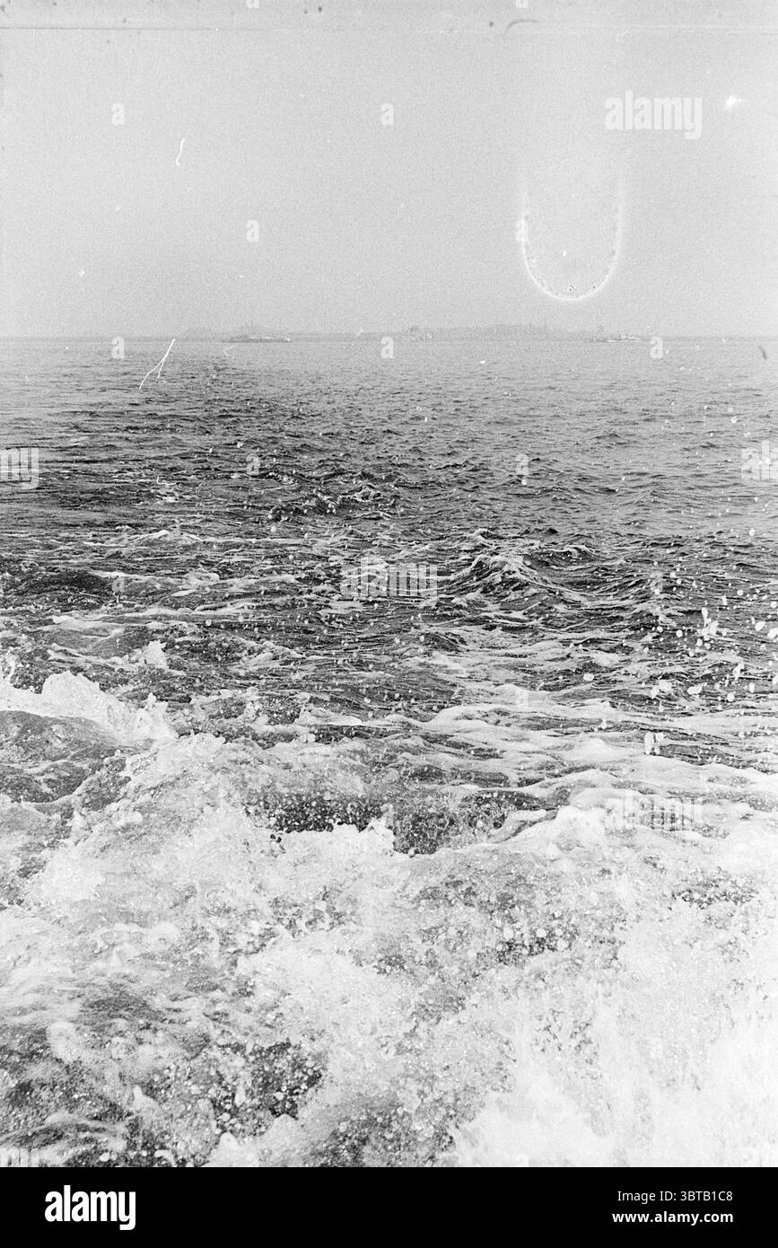 Screw Water., Whizgle News, Dutch Desk, pays-Bas, 1950 - 2000. Ce sont les éléments de l'image. La scène capture une étendue d'eau sereine, où la surface est marquée par de douces vagues qui brillent de divers reflets subtils. L'eau semble s'étirer sans fin vers l'horizon, se mélangeant doucement avec le ciel au-dessus. La palette de couleurs est principalement atténuée, avec des nuances de gris et de bleu profond, lui donnant une sensation tranquille mais un peu monochrome. Au premier plan, les vagues montent et descendent, créant des textures qui évoquent une sensation de mouvement. La mousse blanche de l'eau whe Banque D'Images