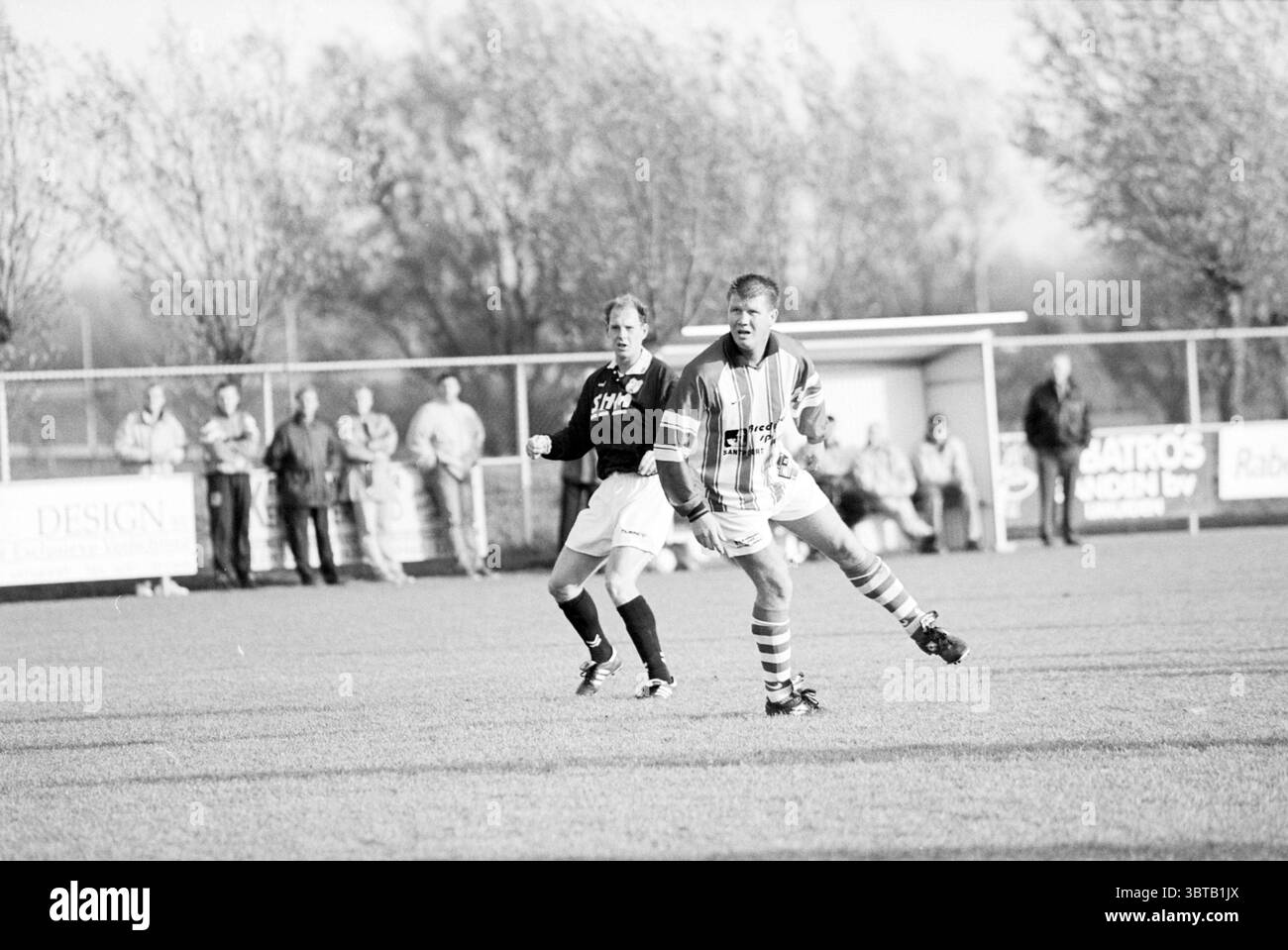 Football VSV - Waterloo, Whizgle News, Dutch Desk, pays-Bas, 1950 - 2000 le 08-11-1997. Ces rubriques sont illustrées dans l'image. La scène présente un match de football animé se déroulant sur un terrain herbeux. Au premier plan, deux joueurs sont en évidence. Un athlète, portant une chemise rayée avec un motif rouge et blanc vif, est engagé dans un mouvement dynamique, se préparant peut-être à frapper le ballon. Sa position reflète la concentration et la détermination, avec un pied légèrement relevé, prêt à l'action. Le deuxième joueur, vêtu d'un maillot noir Uni, semble suivre de près la première pla Banque D'Images