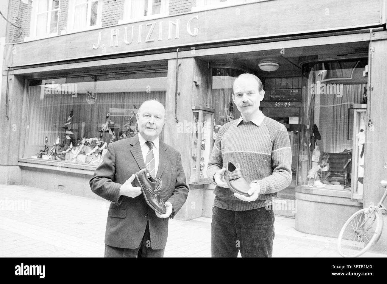 Père et fils Huizing devant leur magasin de chaussures à Grote Houtstraat 42-44 magasins de chaussures et kiosques Haarlem Grote Houtstraat pays-Bas, Whizgle News, Dutch Desk, pays-Bas, 1950 - 2000 le 09-04-1984. L'image inclut ces rubriques. Dans un cadre urbain pittoresque, deux personnes se tiennent bien en vue devant un magasin qui respire une atmosphère charmante et ancienne. La devanture dispose de grandes fenêtres en verre encadrées par des panneaux de bois, présentant une gamme de chaussures élégamment agencées, insistant sur un fort accent mis sur l'artisanat. La première personne, à gauche, est un homme plus âgé avec un di Banque D'Images