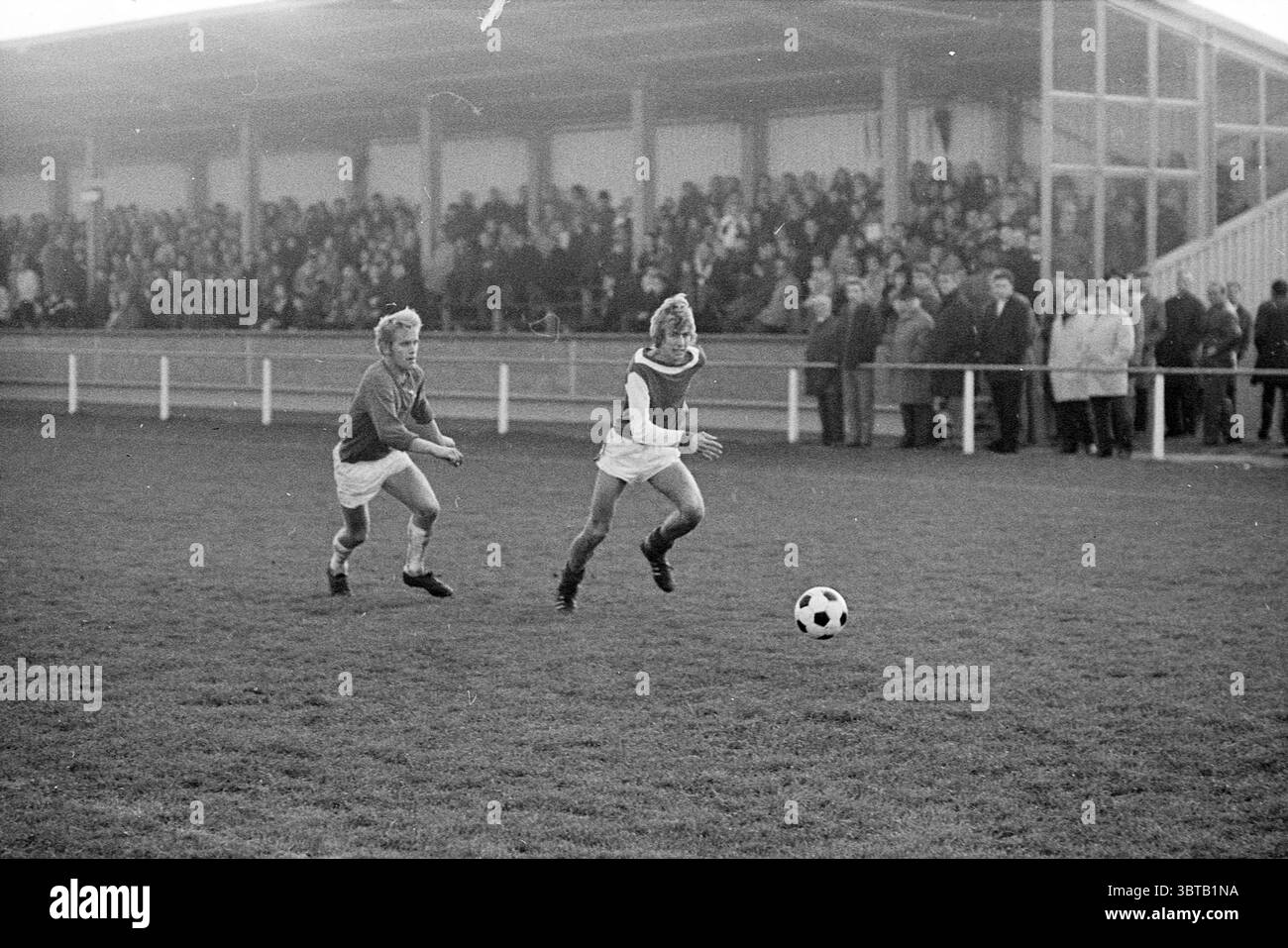 Stormvogels - JOS Football V.S.V. Velsen Stormvogels, Whizgle News, Dutch Desk, pays-Bas, 1950 - 2000 le 09-11-1969. Ces rubriques apparaissent dans l'image. La scène capture un moment dynamique lors d'un match de football tenu dans un cadre animé. Au premier plan, deux jeunes joueurs sont activement engagés dans la poursuite d'un ballon de football. Un joueur, aux cheveux courts et clairs, est légèrement derrière l'autre, qui court devant. Les deux sont vêtus d'uniformes de sport traditionnels : le joueur en tête porte un maillot bleu foncé, tandis que le joueur suivant porte un maillot blanc. Le champ est une alose vibrante Banque D'Images