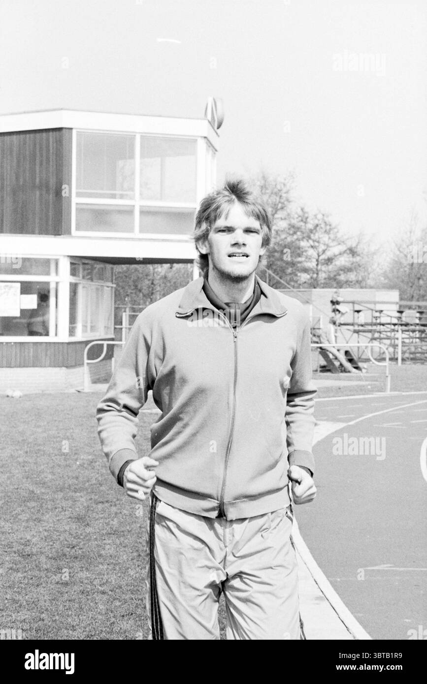 Athlète de course à pied depuis environ 25 ans dans le domaine de l'athlétisme., Whizgle News, Dutch Desk, pays-Bas, 1950 - 2000. Voici les rubriques de l'image. Dans la scène, un jeune homme est en évidence, faisant du jogging avec une expression déterminée sur son visage. Il porte un survêtement de couleur claire, qui se compose d'une veste zippée et d'un pantalon assorti orné de rayures sur les côtés. Le tissu semble léger, reflétant un style sportif décontracté. Le fond révèle un bâtiment moderne avec un style architectural distinctif, présentant de grandes fenêtres qui suggèrent un design contemporain. Surround Banque D'Images