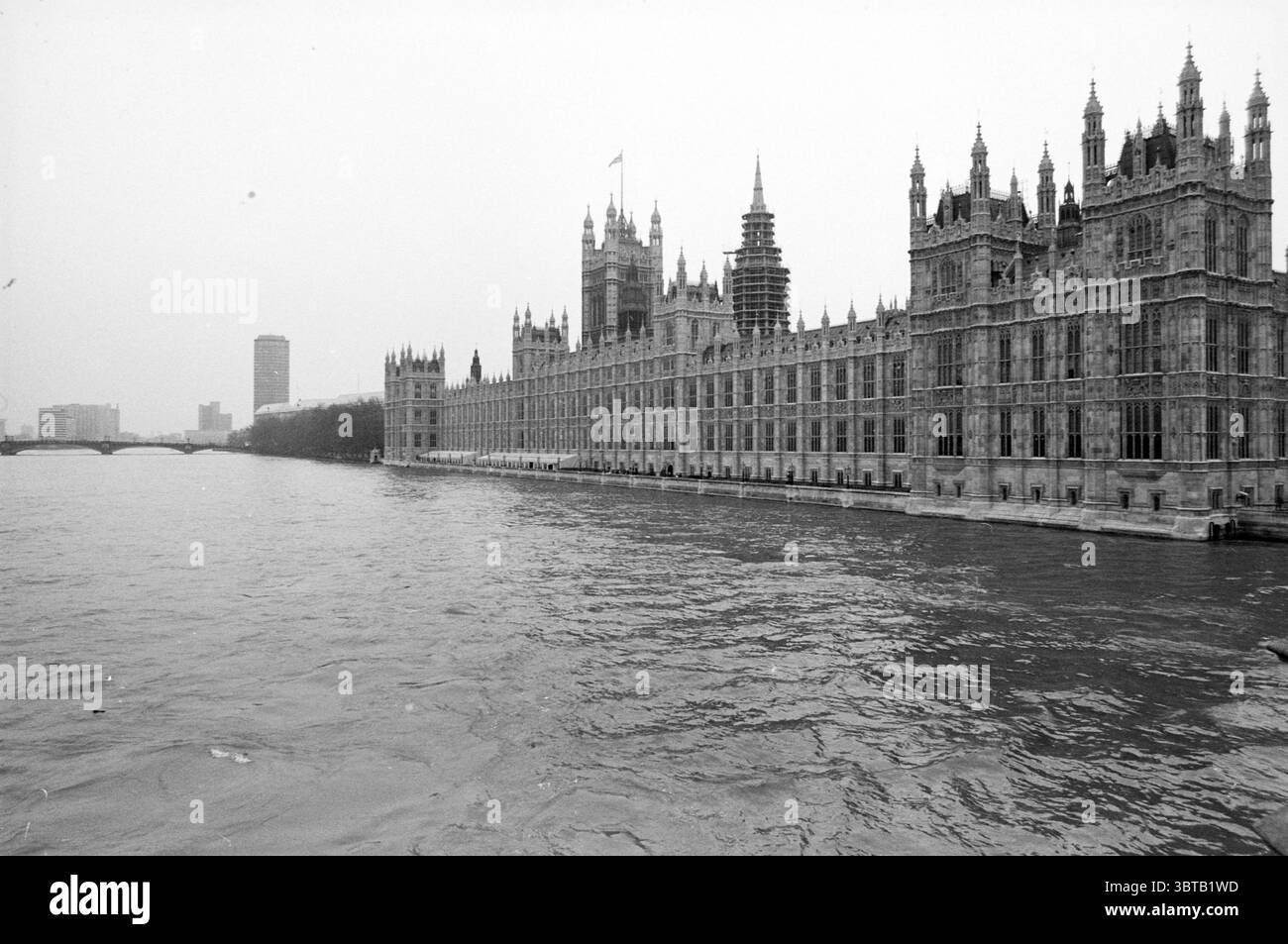 Divers paysages urbains Londres Angleterre Londen Verenigd Koninkrijk, Whizgle News, Dutch Desk, pays-Bas, 1950 - 2000 on 19-04-1988. Ce sont les éléments de l'image. La scène capture une vue grandiose d'un bâtiment emblématique le long de la rivière. Majestueuse dans son architecture, la structure présente des pierres complexes avec de nombreuses flèches pointues et des détails élaborés. Sa présence imposante s'étend le long du bord de l'eau, créant une silhouette saisissante sur le ciel. Le temps semble couvert, avec une teinte grisâtre enveloppant la scène, prêtant une atmosphère sombre mais dramatique. L'eau est Banque D'Images