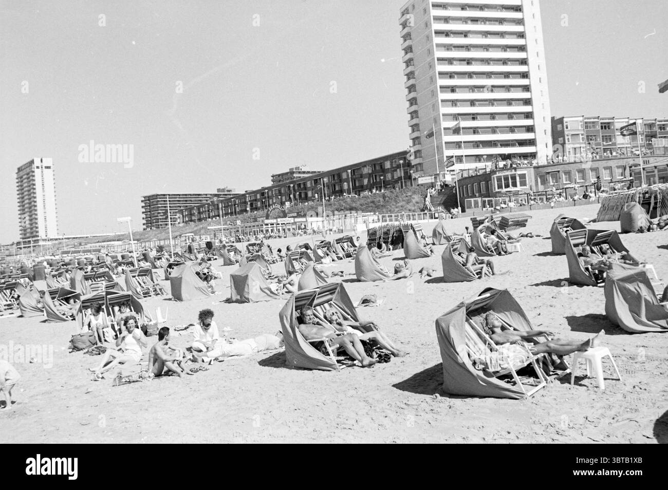 Plage animée Zandvoort plage et plages Zandvoort, Whizgle News, Dutch Desk, pays-Bas, 1950 - 2000 on 28-08-1985. L'image contient ces rubriques. La scène capture une atmosphère de plage animée par une journée ensoleillée. L'étendue sablonneuse est parsemée de nombreuses cabanes de plage, chacune présentant un tissu rayé rouge vif et blanc qui crée un contraste joyeux avec le sable beige pâle. Certaines cabanes sont occupées par des bains de soleil se prélassant tranquillement, tandis que d'autres semblent vides, attendant les visiteurs. Une foule diversifiée peut être vu profiter de la plage ; les gens de différents âges sont étalés sur des serviettes ou nichés Banque D'Images