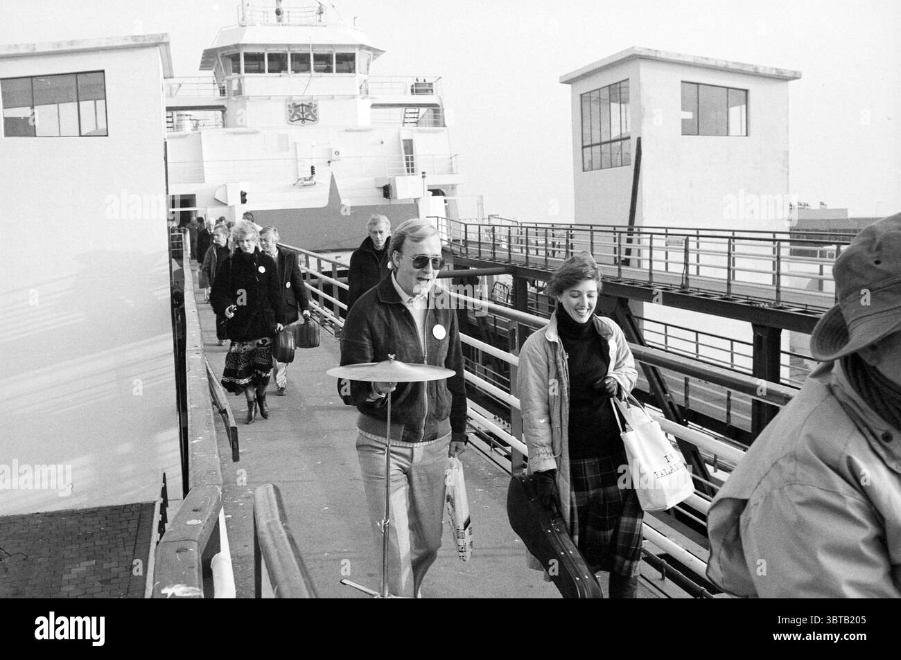 NPO Texel Boat concert North Holland Philharmonic Orchestra, Whizgle News, Dutch Desk, pays-Bas, 1950 - 2000 on 06-11-1982. L'image contient ces rubriques. La scène représente un terminal de ferry animé, où un groupe de personnes débarque d'un grand navire. Le ferry, qui a une structure robuste avec plusieurs ponts, accueille les spectateurs avec un design épuré et net, mettant l'accent sur sa fonction de mode de transport à travers l'eau. Au premier plan, un homme marche avec confiance, tenant avec désinvolture une cymbale aux couleurs vives dans une main, tandis que son autre main agrippe un petit sac. Il w Banque D'Images