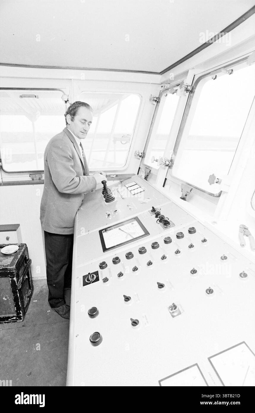 Timonerie de drague, Whizgle News, Dutch Desk, pays-Bas, 1950 - 2000 en 1971. L'image contient ces rubriques. La scène se déroule dans une salle de contrôle, peut-être à bord d'un navire, qui se distingue par une grande console incurvée remplie d'un ensemble de boutons, de commutateurs et de cadrans. Les couleurs du panneau de commande sont principalement atténuées, avec des nuances de gris et de noir, avec quelques accents métalliques qui captent la lumière. Un homme se tient à la barre, s'engageant soigneusement avec les commandes. Il est habillé d'un costume de couleur claire qui contraste avec l'environnement plus sombre, ajoutant une touche de formalité au M. Banque D'Images