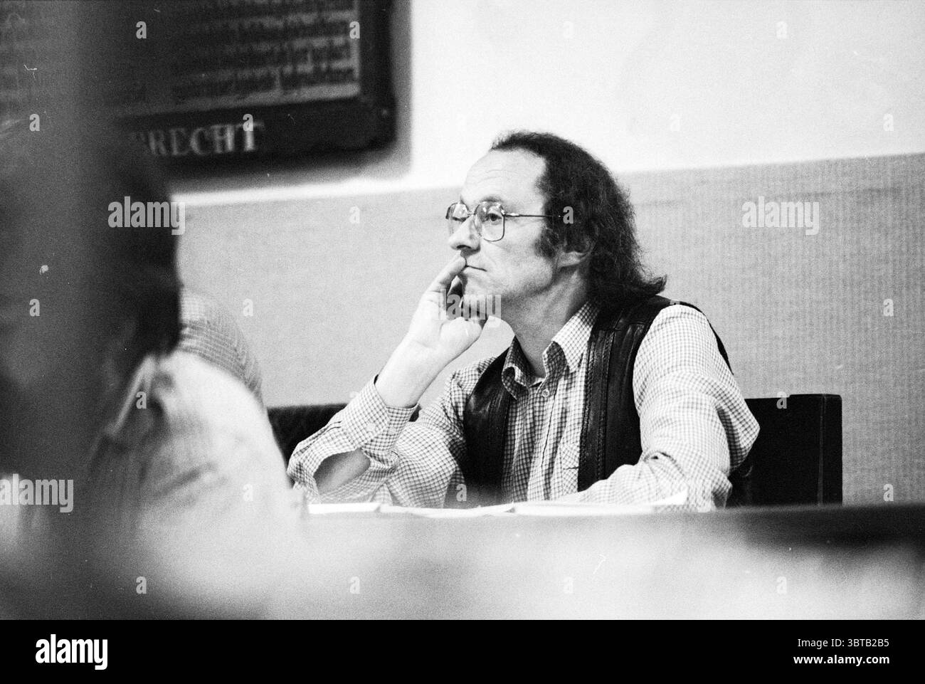 Budget Haarlem Haarlem Conseil municipal Haarlem Grote Markt 2 pays-Bas, Whizgle News, Dutch Desk, pays-Bas, 1950 - 2000 on 02-11-1981. L'image contient ces rubriques. La scène présente un homme assis à une table, profondément dans la pensée. Il a les cheveux longs et foncés, coiffés lâchement, et porte des lunettes qui accentuent son expression contemplative. Sa tenue se compose d'un gilet texturé sur une chemise à carreaux, suggérant un mélange de style décontracté et formel. L'arrière-plan présente un mur beige discret, contribuant à une atmosphère sombre. L'éclairage est doux et diffus, projetant de douces ombres th Banque D'Images