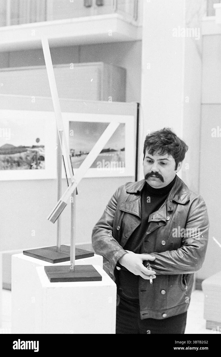 Images d'exposition depuis 100 ans. IJmuiden City Hall Exhibitions, Whizgle News, Dutch Desk, pays-Bas, 1950 - 2000 on 13-02-1976. L'image montre ces rubriques. Dans une galerie spacieuse et bien éclairée, un homme se tient contemplativement à côté d'une sculpture moderne et élégante. La sculpture, caractérisée par ses lignes géométriques et son design minimaliste, se trouve au sommet d'un simple piédestal, attirant l'attention avec sa surface métallique qui reflète la lumière ambiante. L'homme a une moustache sombre et bouclée et est habillé d'une veste en cuir noir, avec un col roulé ajusté en dessous. Il se tient debout les bras croisés, son corps légèrement Banque D'Images