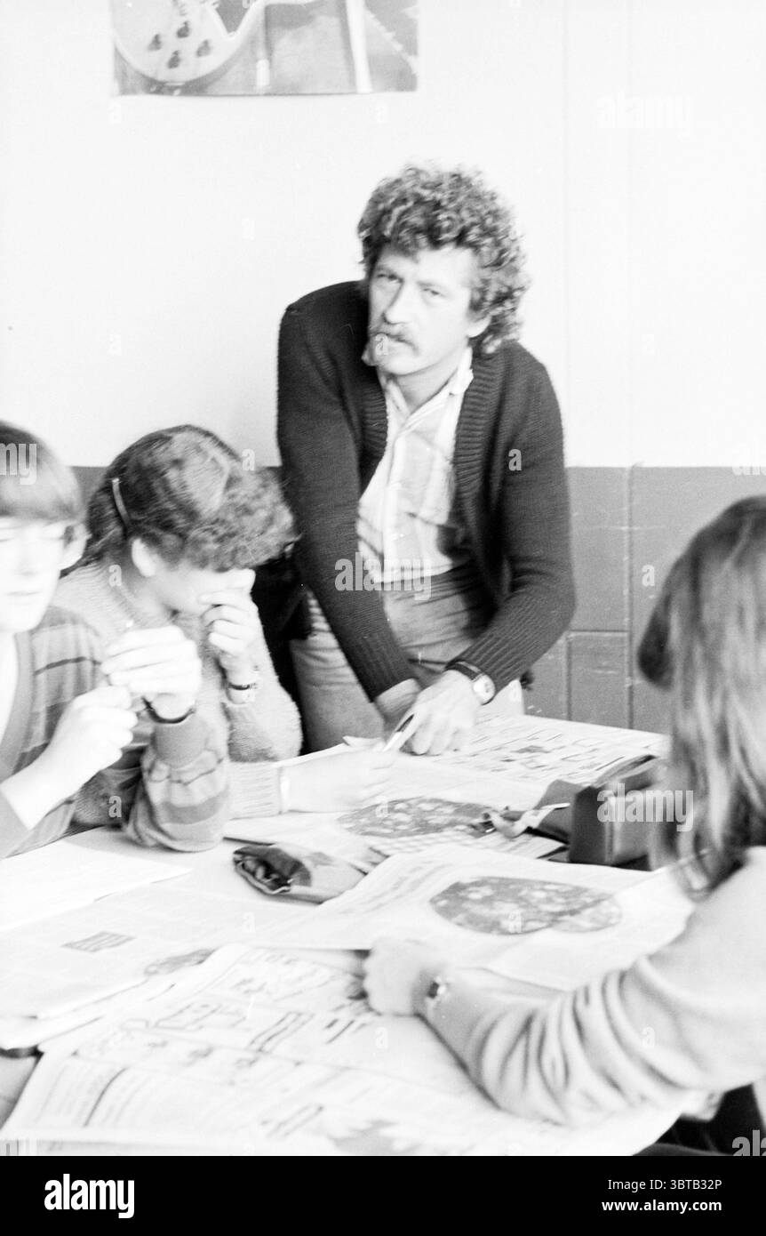 Leçon sur le budget Sassenheim éducation Sassenheim, Whizgle News, Dutch Desk, pays-Bas, 1950 - 2000 le 30-09-1980. L'image contient ces rubriques. Dans un cadre intérieur faiblement éclairé, un groupe d’individus est rassemblé autour d’une table couverte de journaux et de documents divers. L'ambiance semble sérieuse et concentrée, indiquant une atmosphère de collaboration ou de discussion. La figure centrale se tient légèrement penchée, dégageant un sentiment d'autorité ou d'engagement. Il a les cheveux bouclés et une expression faciale qui suggère la concentration comme il se penche sur la table, pointant vers quelque chose avec int Banque D'Images