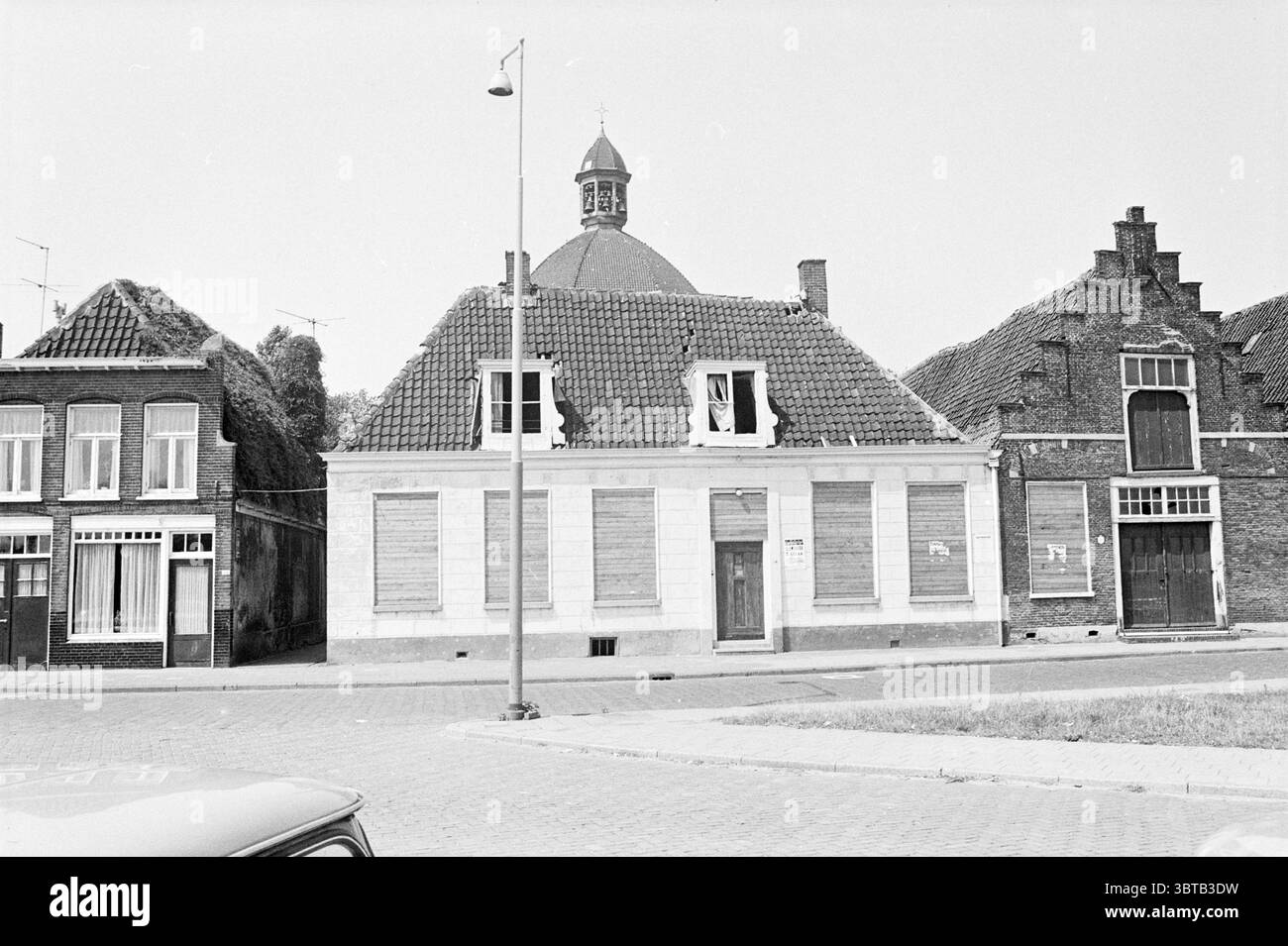 Bâtiments Meerstraat Beverwijk extérieur Beverwijk Meerstraat pays-Bas, Whizgle News, Dutch Desk, pays-Bas, 1950 - 2000 on 07-07-1970. L'image inclut ces rubriques. La scène présente une rue tranquille bordée d'un mélange de bâtiments qui présentent des styles architecturaux distincts. Au premier plan, une grande structure de deux étages avec un dôme proéminent et arrondi se distingue, sa surface marquée par des briques altérées et un toit en ardoise présentant un motif distinctif. Les fenêtres du bâtiment sont arborées, suggérant un abandon ou une rénovation, ce qui donne un sentiment de négligence. À côté de Banque D'Images