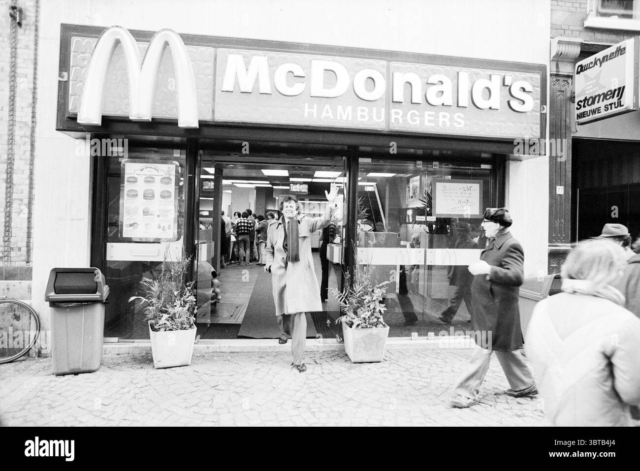 Traiteur Grote Houtstraat traiteur Haarlem Grote Houtstraat pays-Bas, Whizgle News, Dutch Desk, pays-Bas, 1950 - 2000 on 01-03-1982. L'image montre ces rubriques. La scène capture un environnement urbain animé, dominé par la façade proéminente d'un établissement de restauration rapide. Le bâtiment dispose d'une grande entrée accueillante avec une porte vitrée encadrée par des arches jaunes lumineuses et emblématiques au-dessus. L'extérieur est principalement revêtu de tons discrets, ce qui améliore l'esthétique rétro de l'ambiance générale. Devant l'entrée, une figure se distingue, dégageant un air de confiance décontractée. Dre Banque D'Images