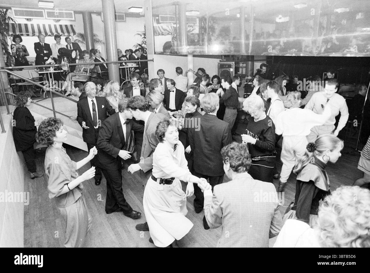 Festiball Concertgebouw Football Haarlem, Whizgle News, Dutch Desk, pays-Bas, 1950 - 2000 le 28-01-1989. L'image contient ces rubriques. La scène est animée et remplie de mouvement, représentant un rassemblement social bondé, probablement une danse ou une célébration. Au premier plan, un groupe diversifié de personnes est engagé dans la danse, échangeant des sourires et des rires. La tenue est formelle, avec de nombreuses personnes vêtues de costumes et de robes élégantes, présentant un mélange de couleurs sombres et claires, ainsi que des motifs tels que des rayures et des fleurs. Certaines tenues ont un style des années 80, avec des épaulettes et des styles audacieux creat Banque D'Images