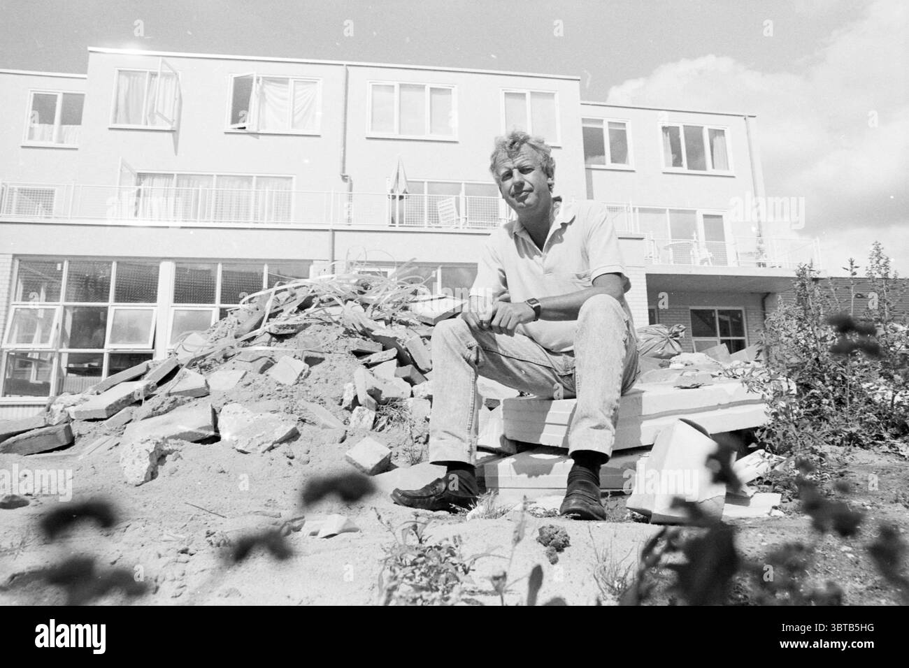 Playground Jan Gijsen School, Whizgle News, Dutch Desk, pays-Bas, 1950 - 2000 le 07-08-1990. L'image contient ces rubriques. Dans la scène, un jeune homme est assis sur un tas de débris, qui se compose de matériaux de construction cassés comme des briques et du plâtre. Il a des cheveux blonds et est habillé d'une chemise à manches courtes de couleur claire jumelée à un pantalon pâle. Sa posture est détendue ; il regarde directement devant lui avec une expression calme. Le fond présente un bâtiment moderne avec de grandes fenêtres, dont certaines sont ornées de rideaux ouverts, révélant un aperçu de l'intérieur. La structure, PA Banque D'Images
