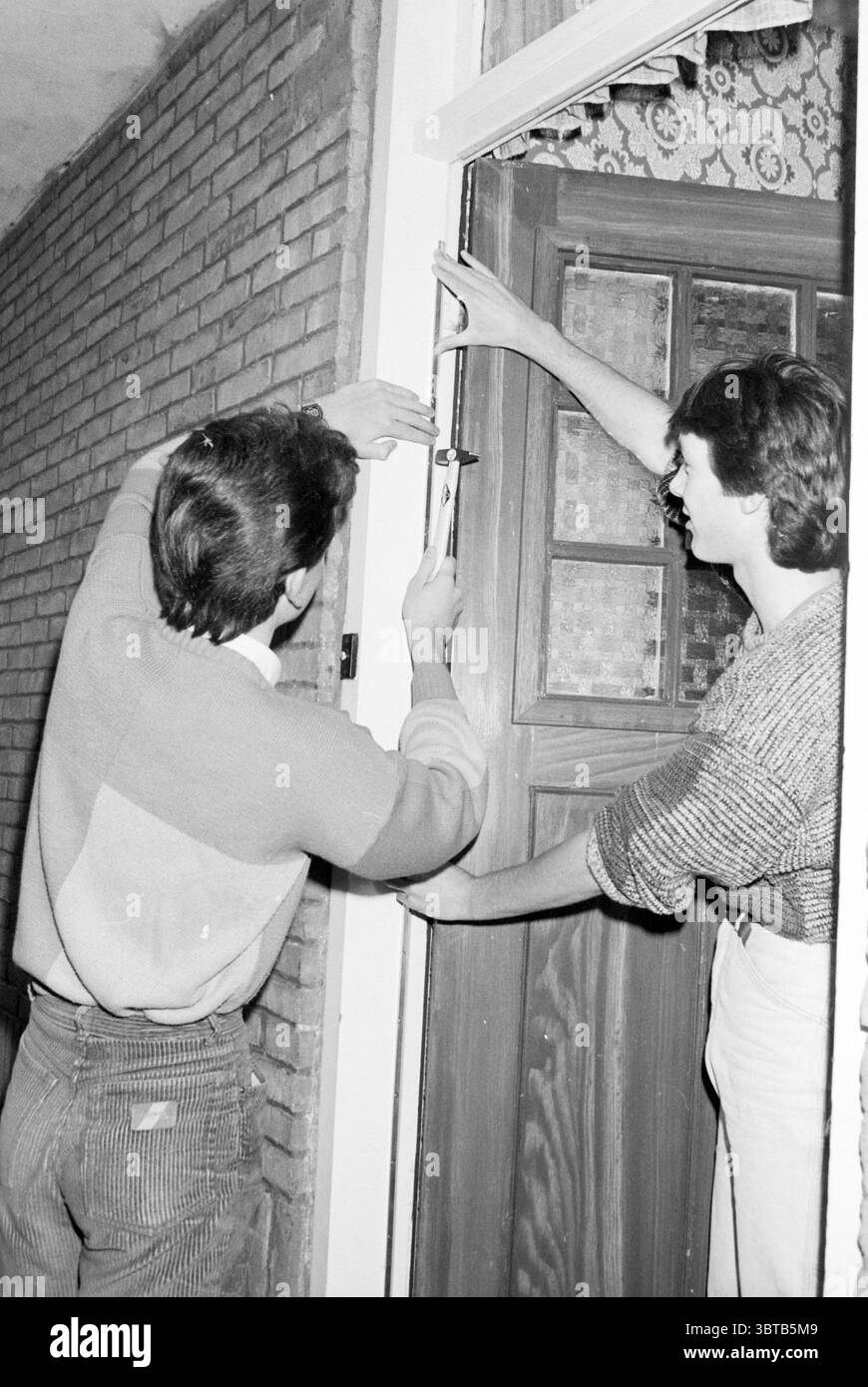 Campagne bricoleur IJmond MTS Schools, Whizgle News, Dutch Desk, pays-Bas, 1950 - 2000 le 21-12-1984. L'image contient ces rubriques. Dans un cadre intérieur confortable, deux individus sont engagés dans une tâche collaborative. Les murs sont ornés d'un motif de briques texturées, prêtant un charme rustique à l'espace. La lumière naturelle filtre à travers une fenêtre voisine, projetant des ombres douces et éclairant la scène d'une lueur chaude. Une personne se tient à gauche, concentrée intensément sur un outil, probablement un appareil de mesure, tenu dans ses mains. Cet individu porte un pull de couleur claire, le tissu apparaissant s Banque D'Images