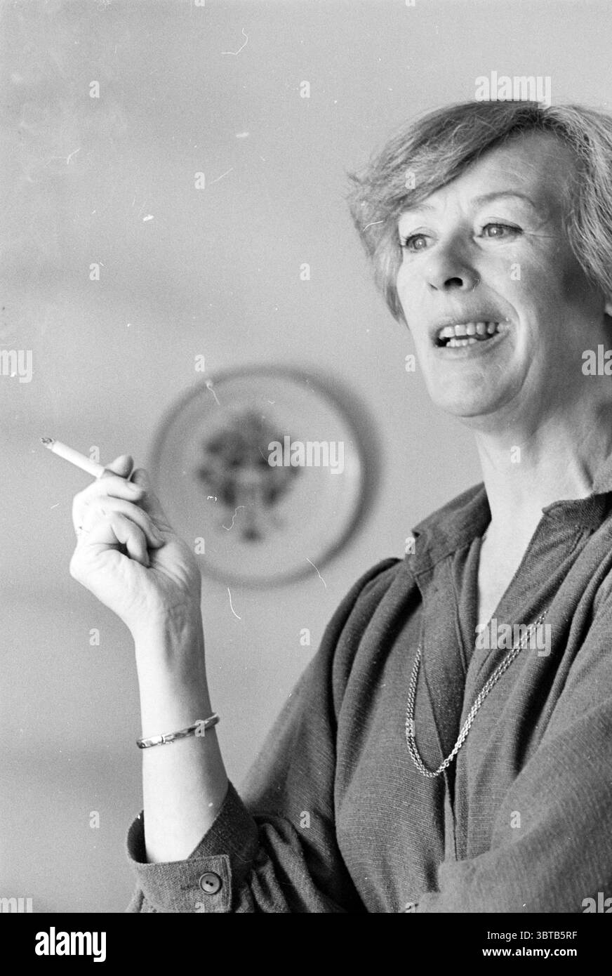 Walking with Elisabeth Andersen Walking, Whizgle News, Dutch Desk, pays-Bas, 1950 - 2000 on 23-04-1978. Voici les rubriques de l'image. Dans la scène, une femme se tient dans un espace bien éclairé qui suggère une atmosphère calme et intime. Elle a une posture confiante, avec son corps légèrement incliné lorsqu'elle engage la conversation. Ses cheveux sont coiffés d'une manière douce et sophistiquée, tombant gracieusement autour de son visage. La femme tient délicatement une cigarette entre ses doigts, la fumée s'enroulant doucement dans l'air, faisant allusion à un moment détendu ou à une pensée profonde. Elle porte un simple mais élégant Banque D'Images