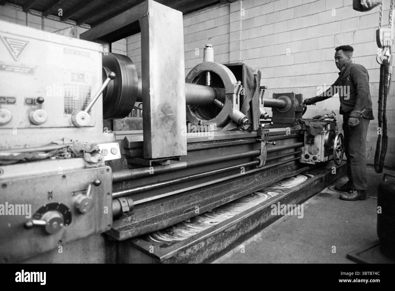 Mr. Leek et Dol Ned. Fonderies de métaux, Whizgle News, Dutch Desk, pays-Bas, 1950 - 2000 sur 26-12-1995. L'image contient ces rubriques. La scène capture un atelier rempli d’une atmosphère industrielle. Au premier plan, une grande machine de tour occupe le devant de la scène, faite de métal robuste avec une surface légèrement usée, indiquant son utilisation intensive. La machine est dominée par une pièce cylindrique qui semble être en cours de mise en forme, avec divers cadrans et leviers visibles sur son côté, faisant allusion à sa fonction technique. Sur le côté, un homme se tient attentif au tour. Lui Banque D'Images