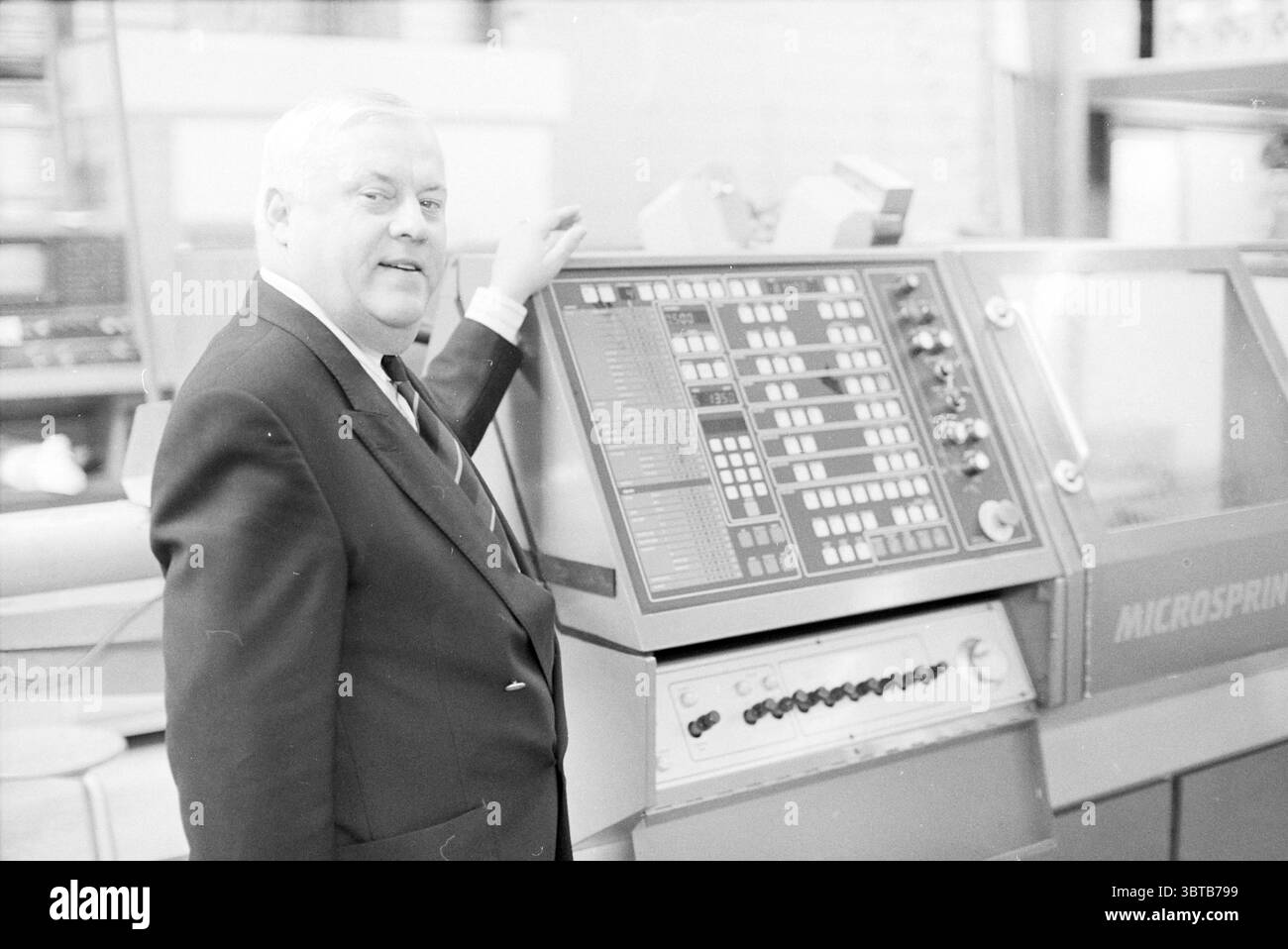 Machine Micosprint 50 pour tournage automatisé, Whizgle News, Dutch Desk, pays-Bas, 1950 - 2000 sur 10-1991. L'image contient ces rubriques. La scène montre un monsieur plus âgé debout à côté d'un panneau de commande complexe. Il est habillé d'un costume sombre formel, donnant un sens du professionnalisme et de l'autorité. Ses cheveux sont courts et blancs, contrastant fortement avec le tissu plus foncé de son costume. Il a un sourire confiant, suggérant un sentiment de fierté ou de satisfaction dans ce qu'il démontre. Le panneau de commande lui-même est un élément important, couvert par une variété de boutons, cadrans et numériques Banque D'Images