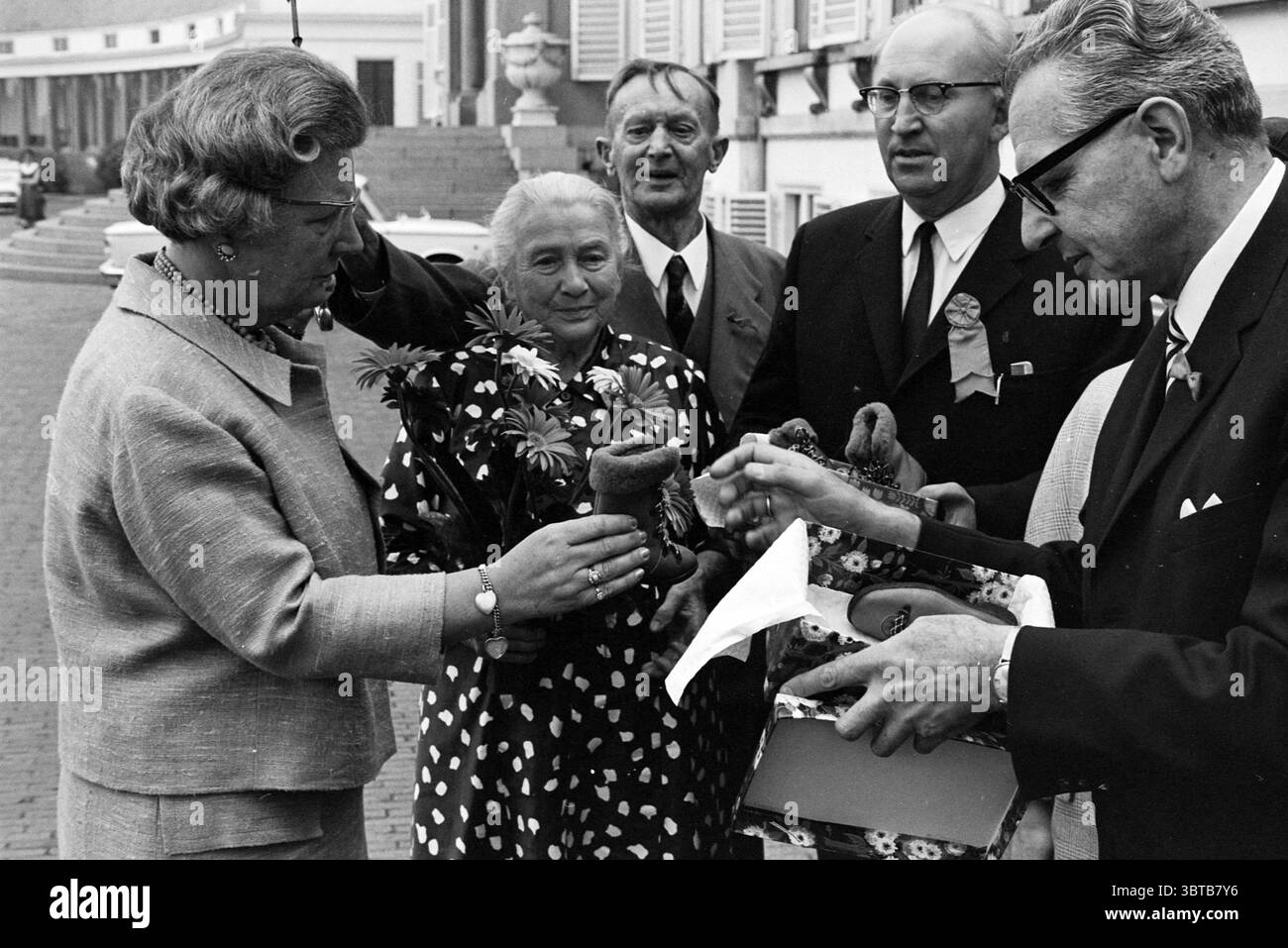 Défilé des personnes âgées Soestdijk Palace Baarn réceptions royales et visites royales Baarn Amsterdamsestraatweg 1, Whizgle News, Dutch Desk, pays-Bas, 1950 - 2000 on 18-06-1968. L'image inclut ces rubriques. Dans un cadre extérieur formel, un groupe de cinq personnes est engagé dans une interaction animée. La scène se caractérise par un mélange de tons doux et neutres, évoquant un sentiment de nostalgie. Au premier plan, une femme, vêtue d'un costume de couleur claire, respire l'élégance avec ses cheveux stylés et son comportement bien fixé. Elle tient un bouquet de fleurs fraîches, qui comprend des pétales délicats dans les tons de Banque D'Images
